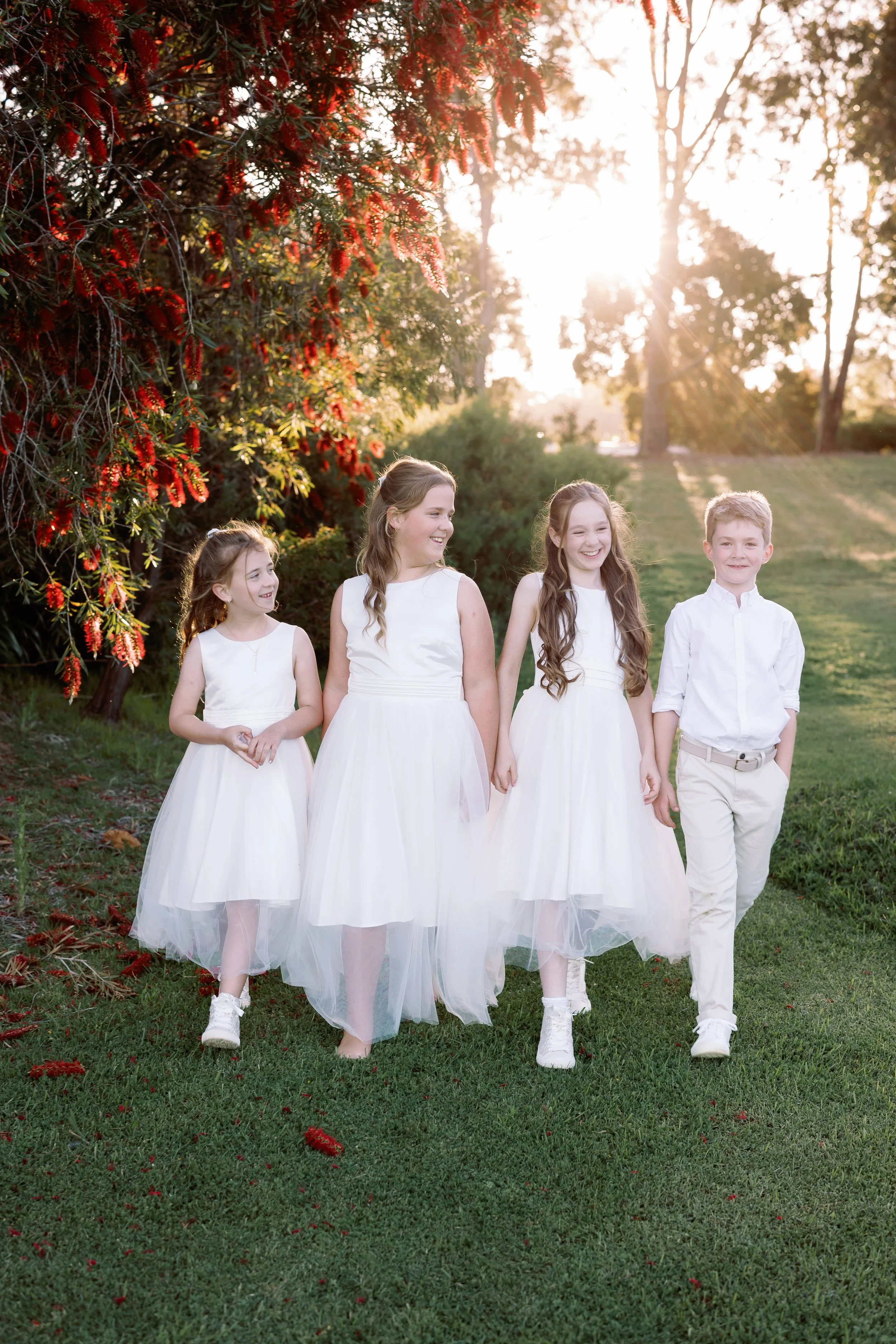 Four children, three girls in white dresses and one boy in white pants and shirt, walking outdoors on grass at sunset, smiling and holding hands.