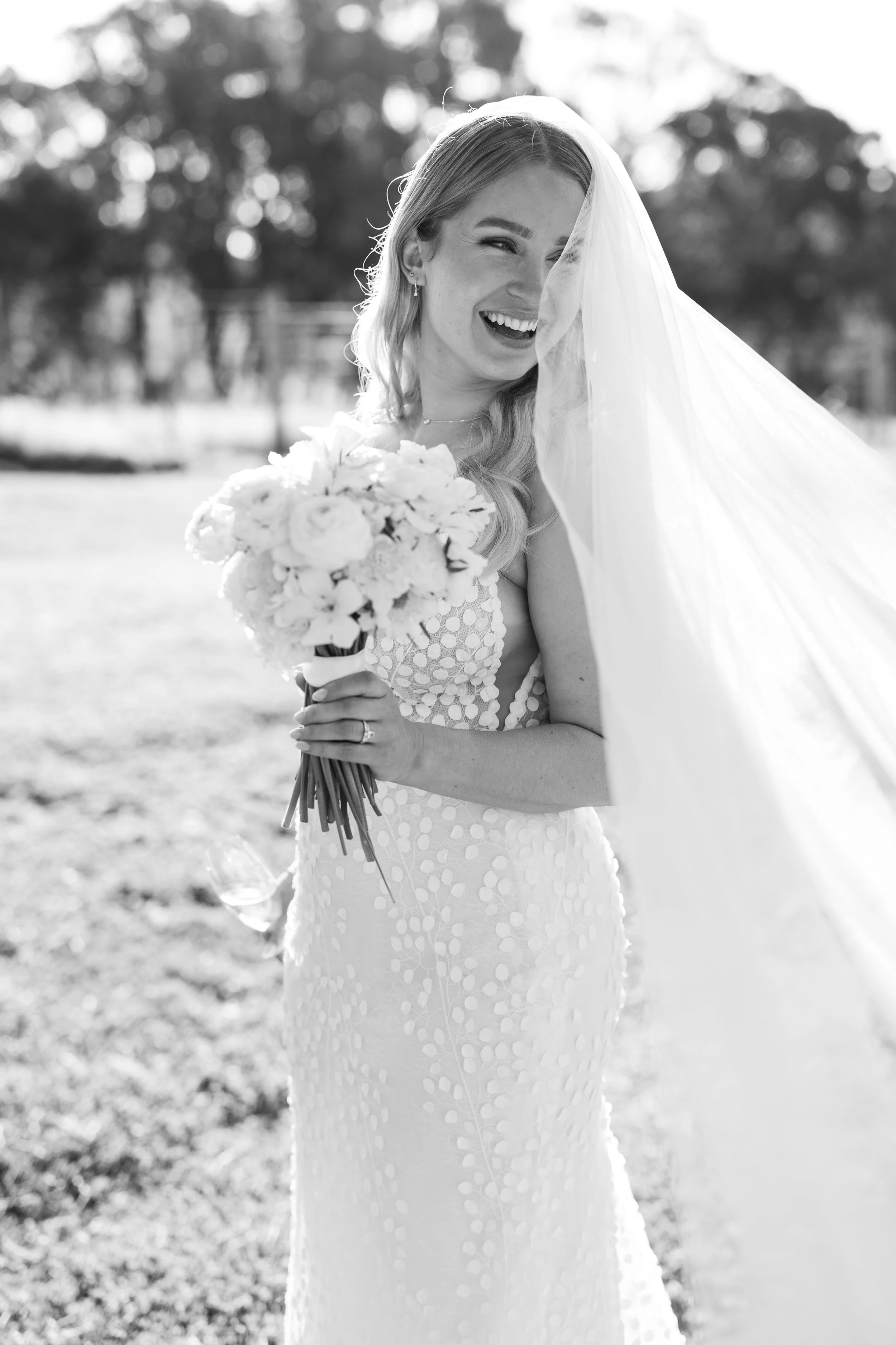 Black and white photo of a smiling bride holding a bouquet of flowers outdoors, wearing a dress with a patterned bodice and a long veil over her face.