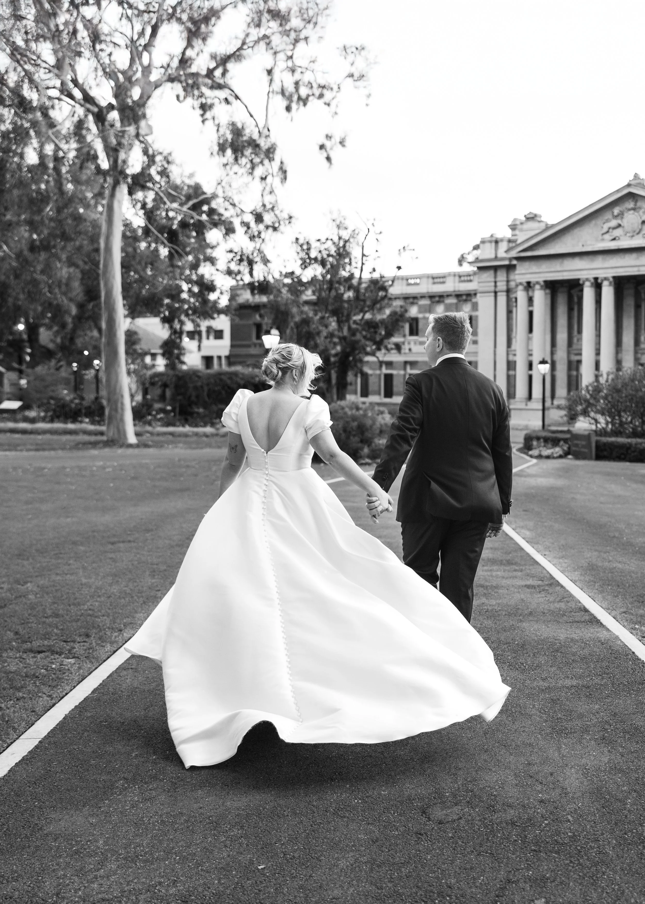 A black-and-white photo of a bride and groom walking hand in hand on a park pathway, with the bride wearing a formal gown and the groom in a suit, in front of a historic building with columns at the State Buildings Perth 