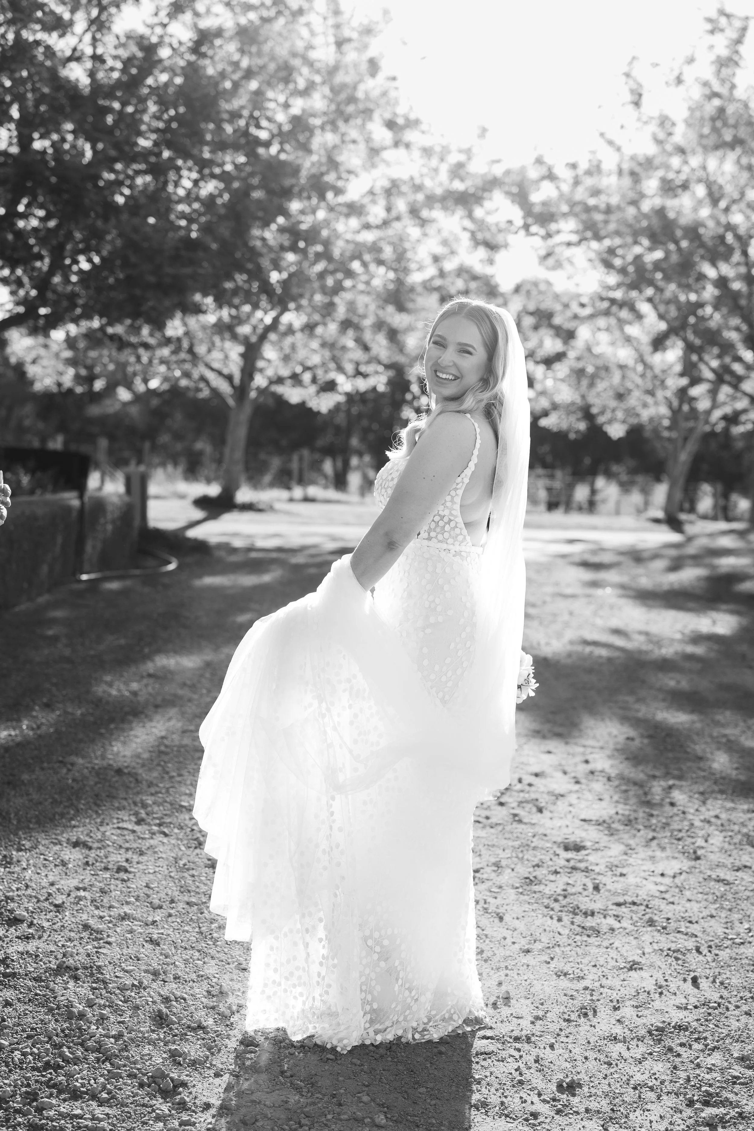 A smiling woman in a wedding dress walking outdoors on a sunny day, trees in the background, captured in black and white.