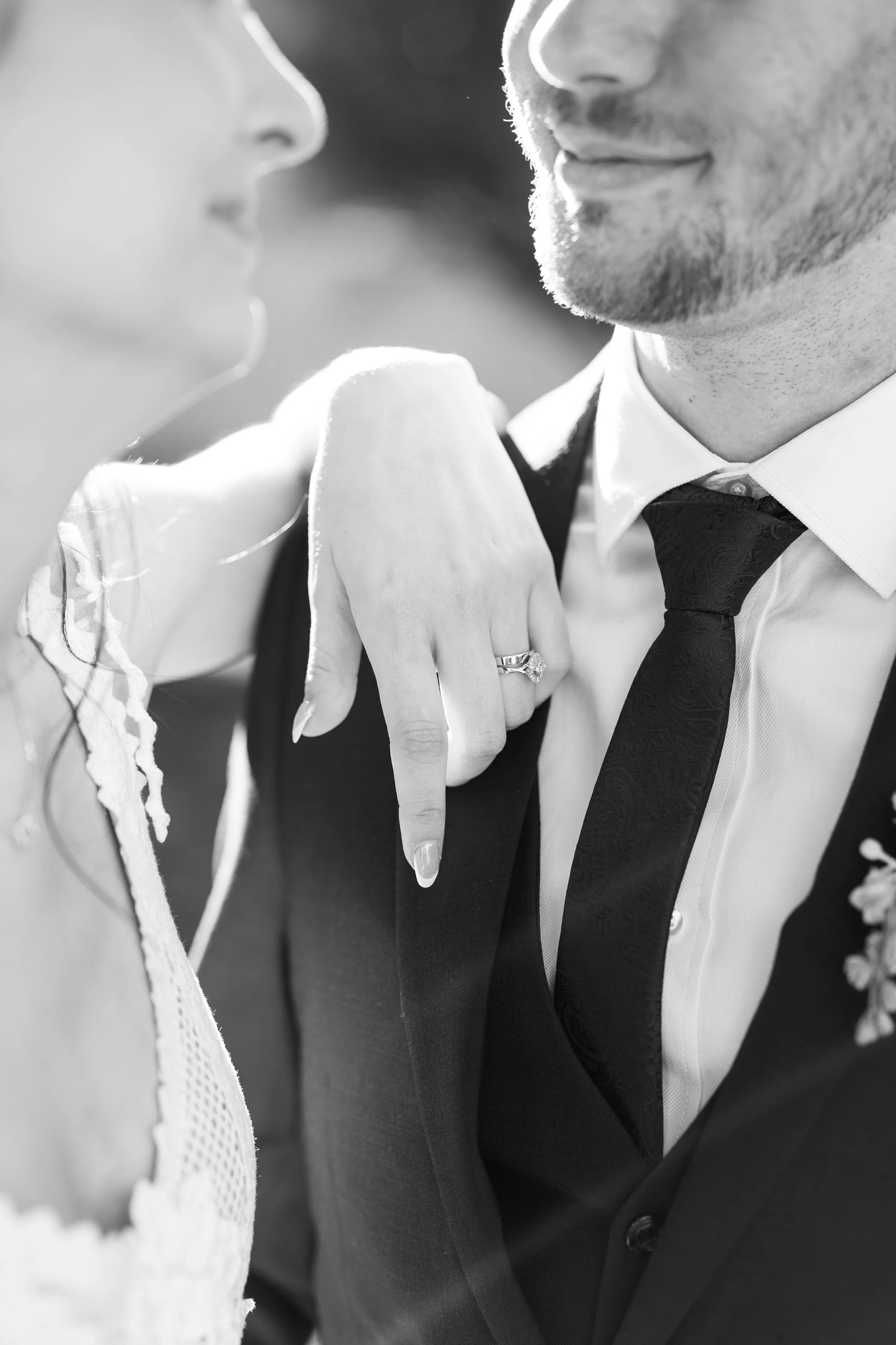 Close-up of a bride and groom showing only their noses, lips, and part of their faces; the bride's hand with an engagement ring resting on the groom's shoulder, in black and white.