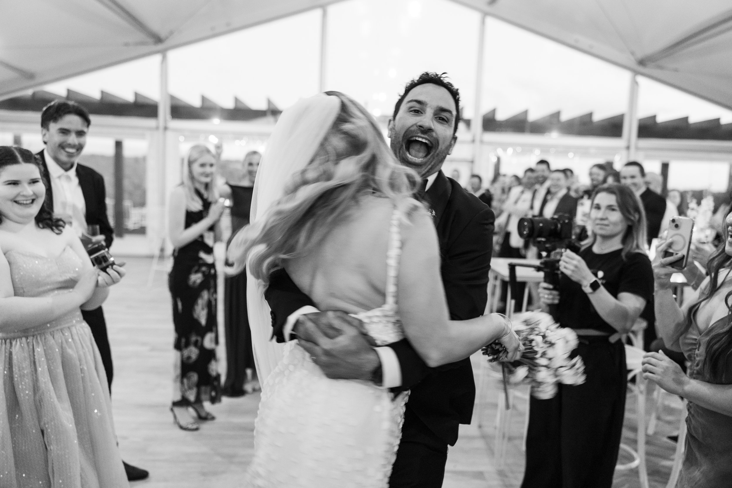 A joyful wedding dance scene with a couple hugging, surrounded by guests capturing the moment, inside a decorated venue.