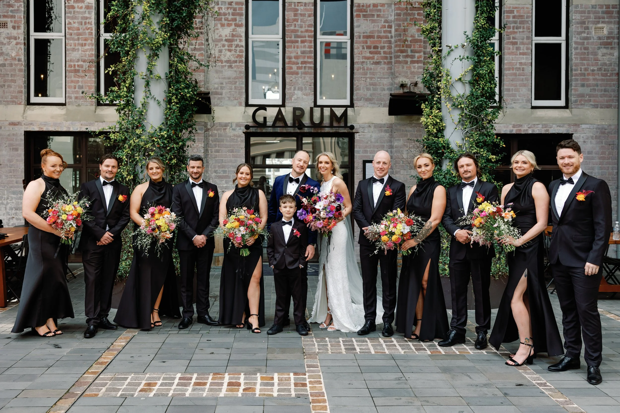 A wedding party standing outside a brick building with the sign garum. The bride and groom are in the center, surrounded by bridesmaids and groomsmen, all dressed in formal attire. The bridesmaids are holding bouquets of colorful flowers, and the gro