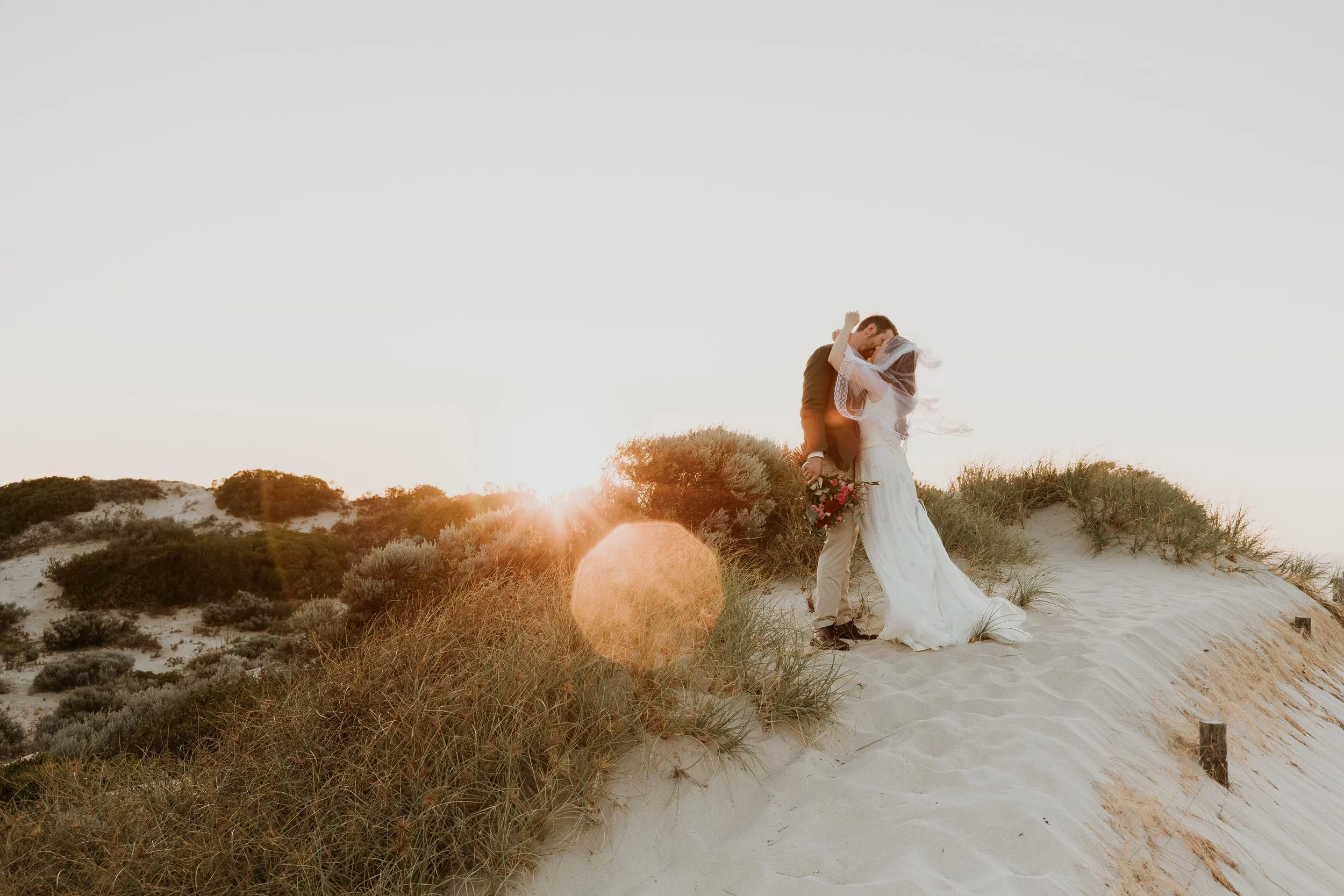 A bride and groom kissing on a sandy dune with grass and bushes during sunset.