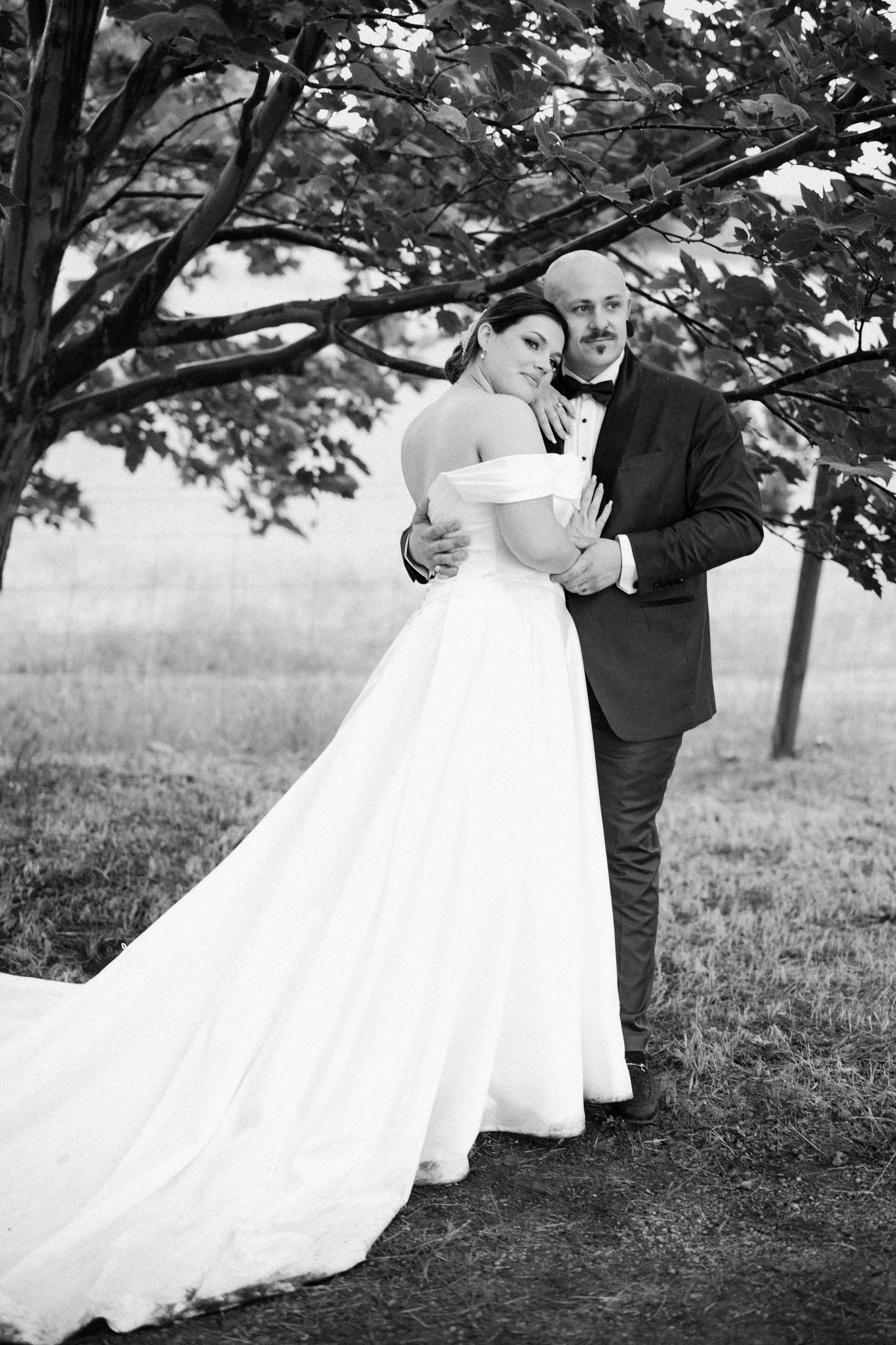 Black and white photograph of a bride in a wedding dress and a groom in a tuxedo standing under a tree, embracing each other and looking at the camera.