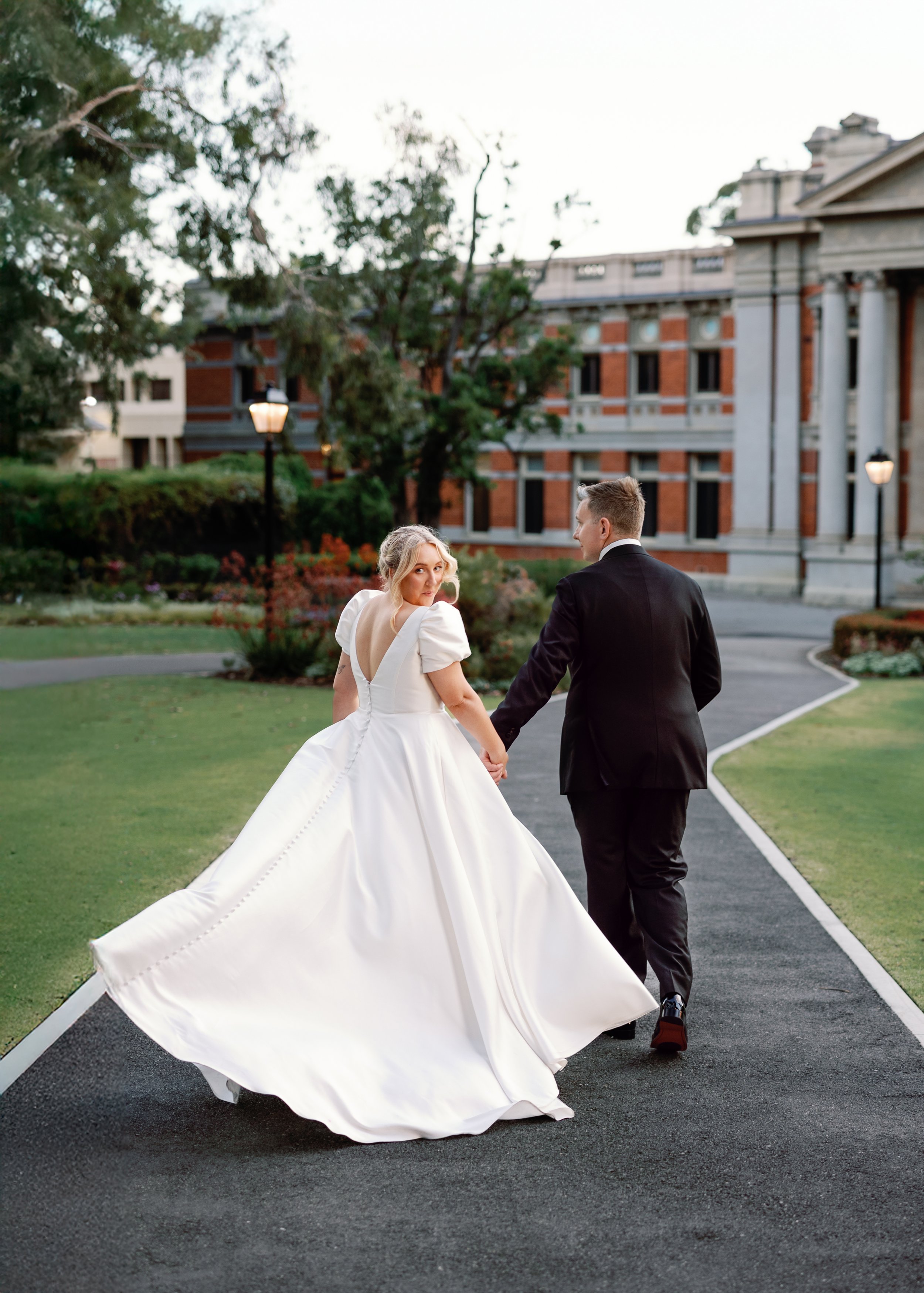 Bride and groom holding hands walking on a paved path outdoors during sunset, with a historic building and trees in the background.