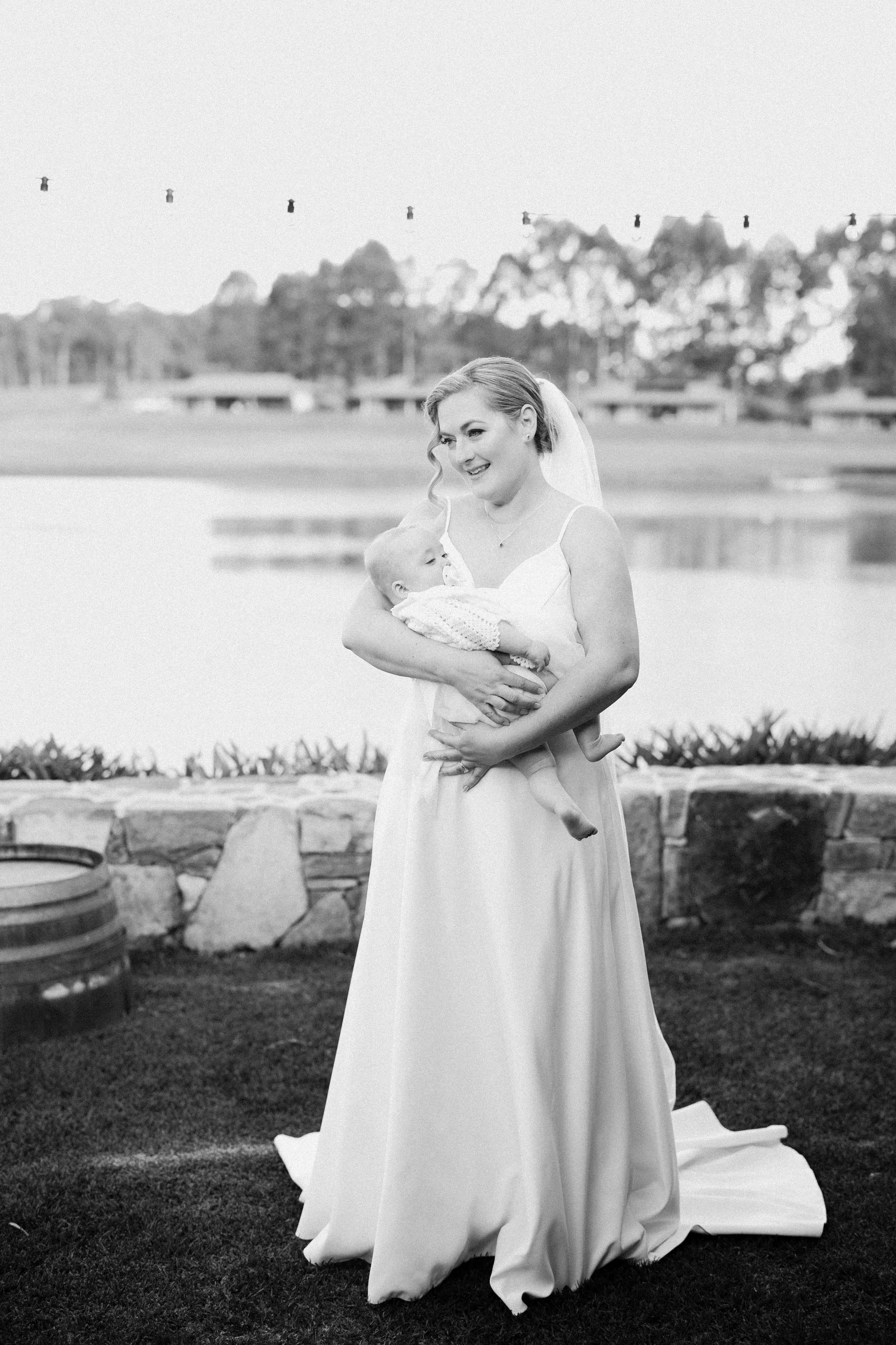 A woman in a wedding dress holding a baby, standing outdoors near a body of water with trees in the background.