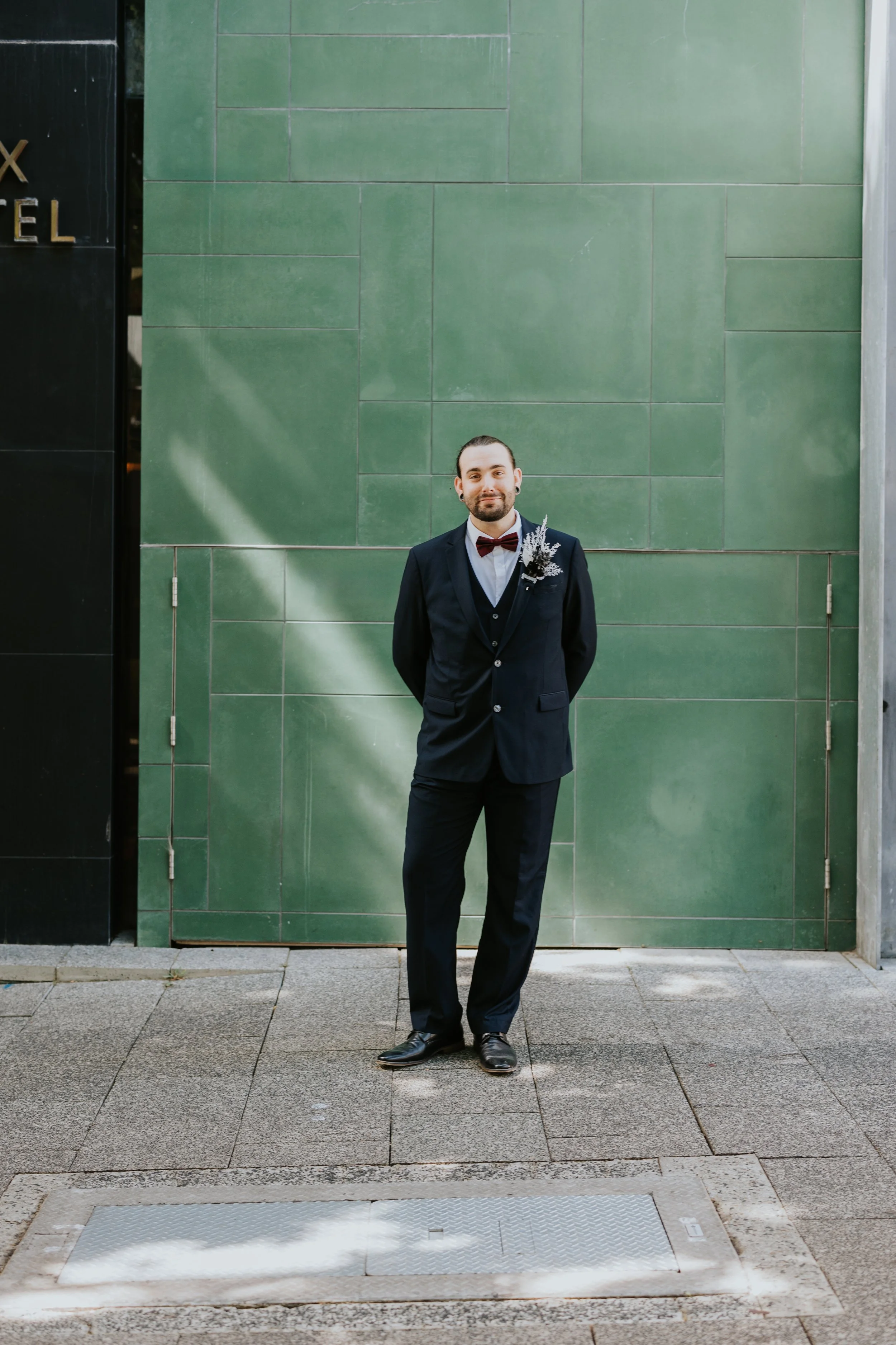 A man dressed in formal suit with a boutonniere, standing outdoors on a paved sidewalk in front of a green tiled wall.