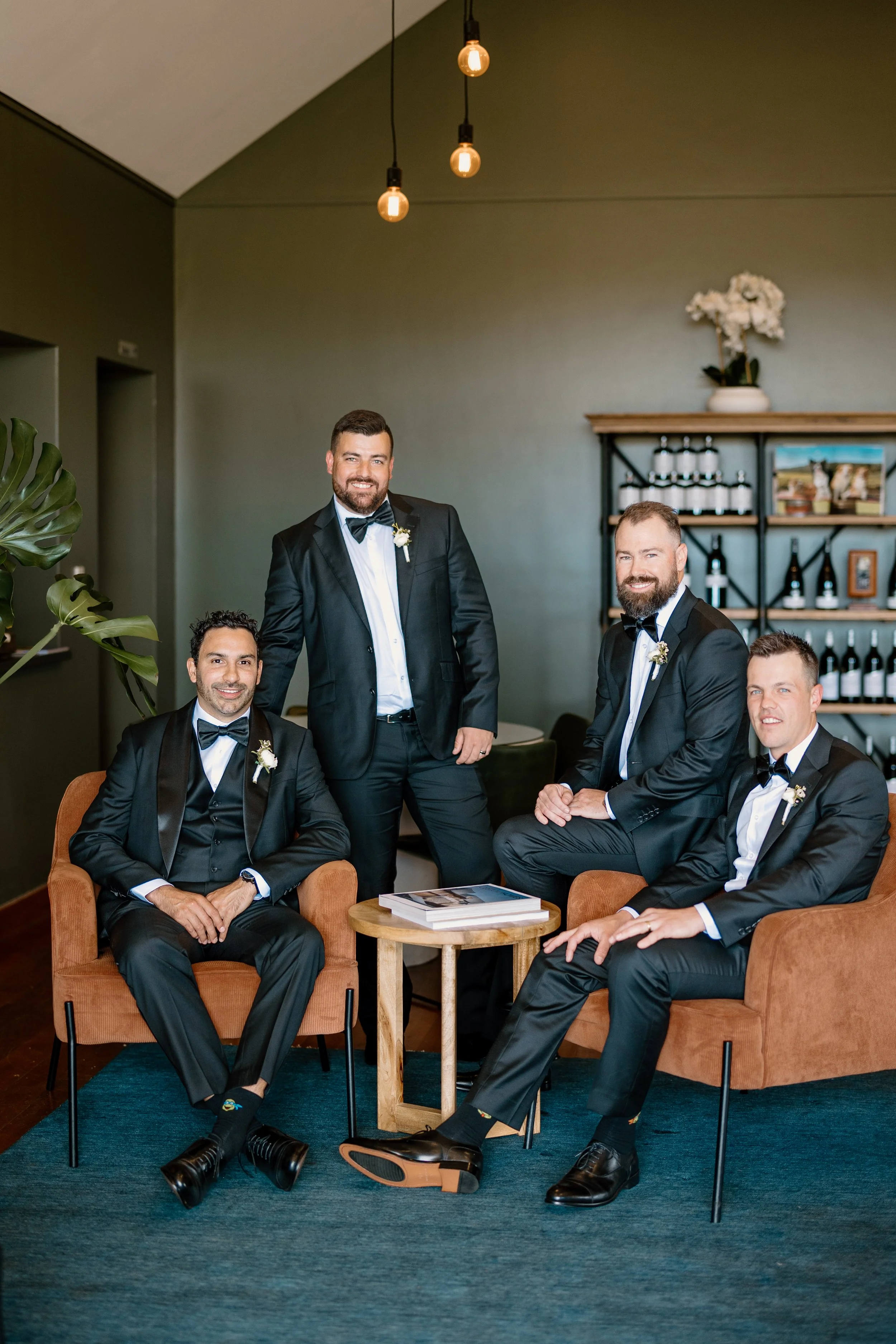 Four men in tuxedos smiling and sitting on brown chairs with a small wooden table between them, in a room with green walls, a bar shelf with wine bottles, and hanging lightbulbs.