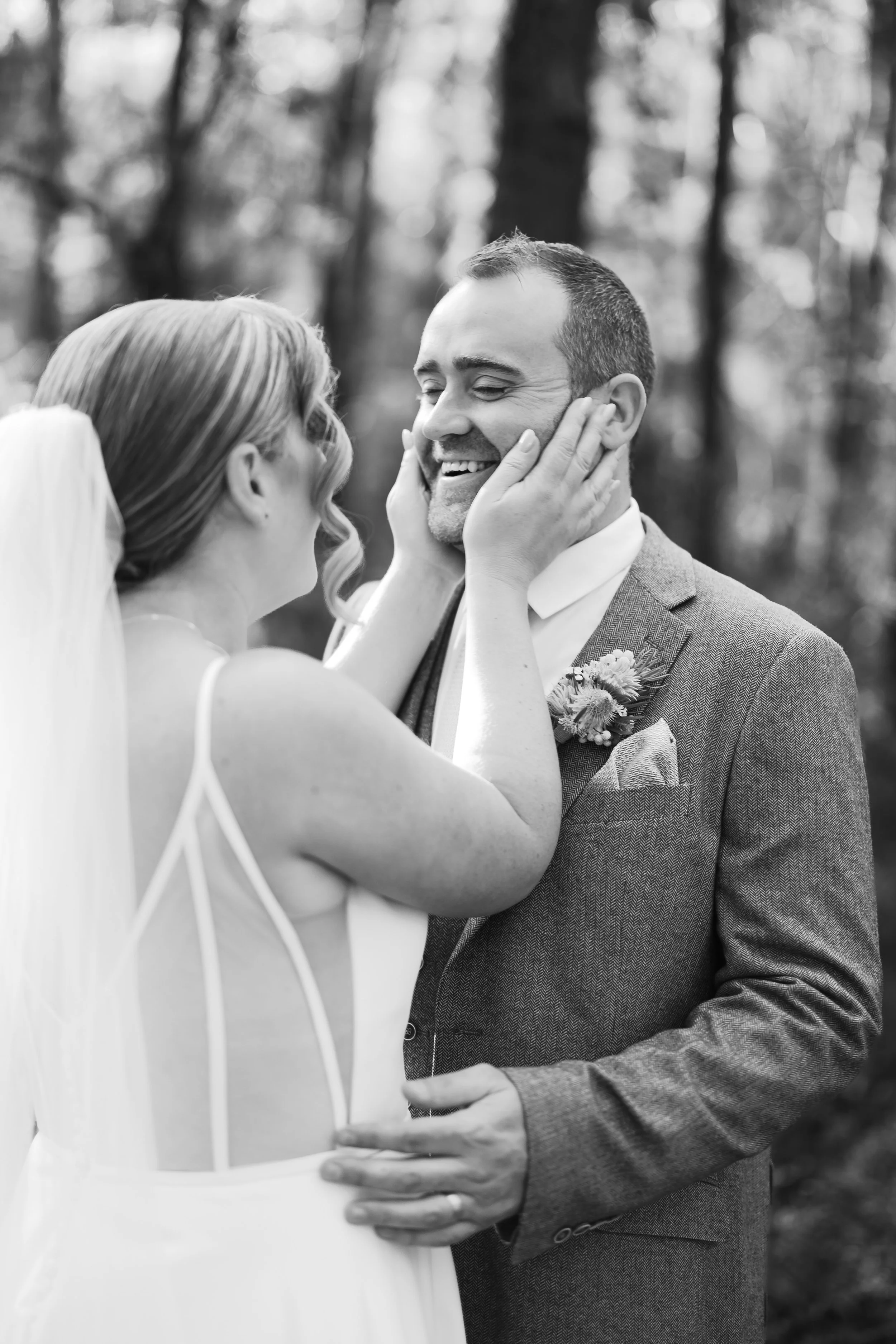 Black and white photo of a bride and groom sharing a joyful moment, with the bride holding the groom's face and both smiling, outdoors with trees in the background.