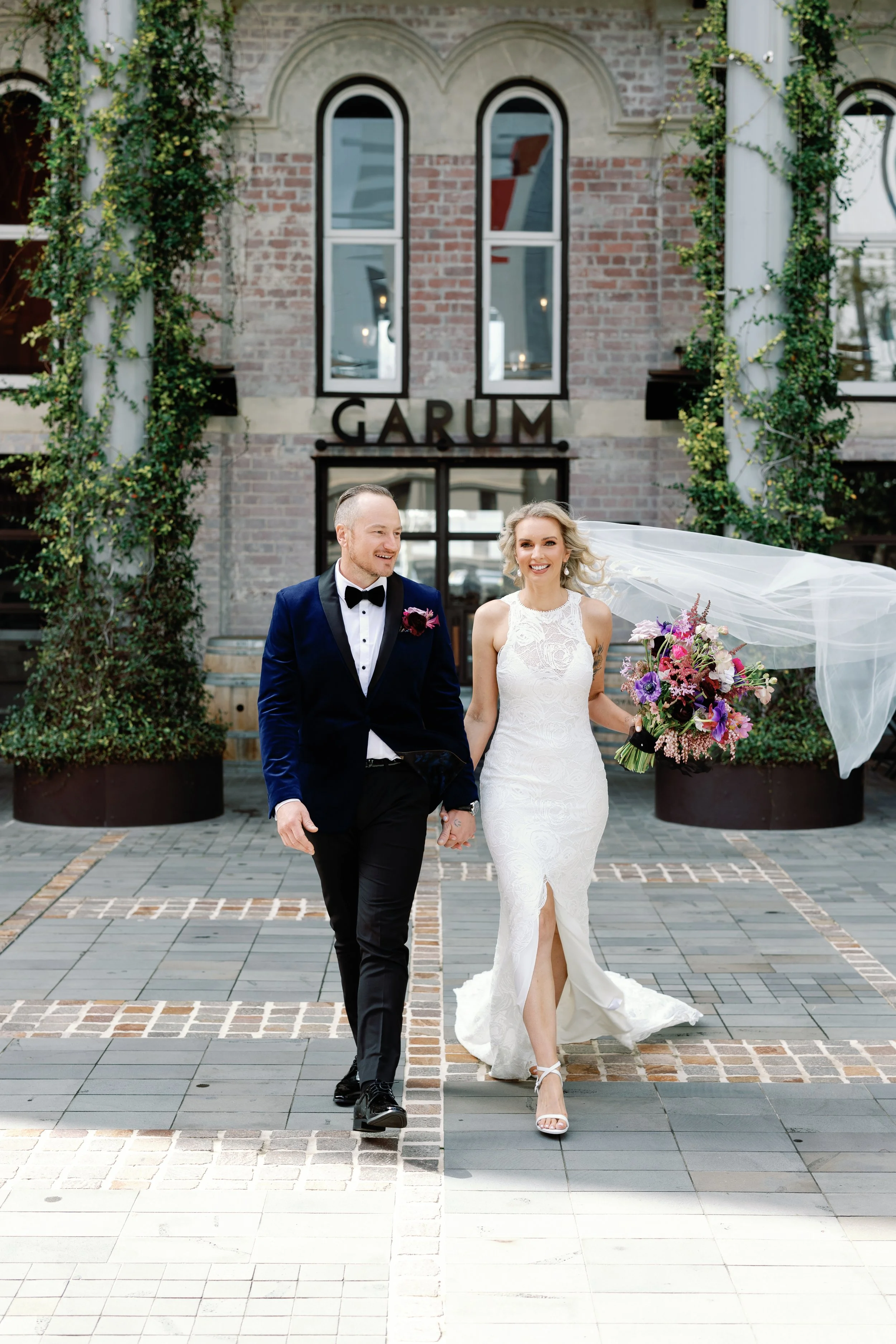 A bride and groom walking hand in hand outside a brick building with large windows and a sign that reads 'GARUM'. The bride is in a white lace wedding dress holding a colorful bouquet, and the groom is in a navy tuxedo with a black bow tie.