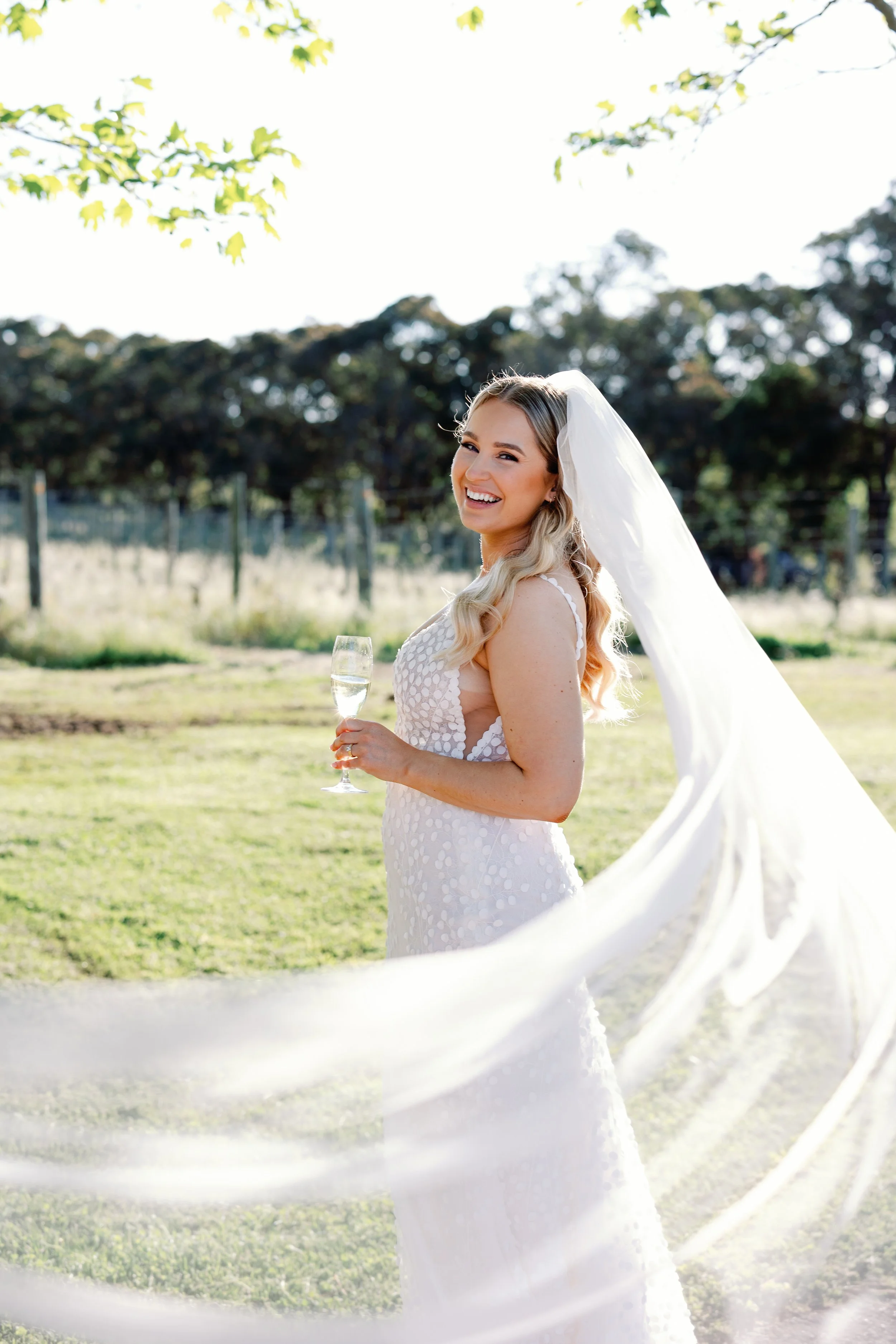 A bride with long blonde hair in a white wedding dress holding a glass of champagne, smiling outdoors with greenery and trees in the background.