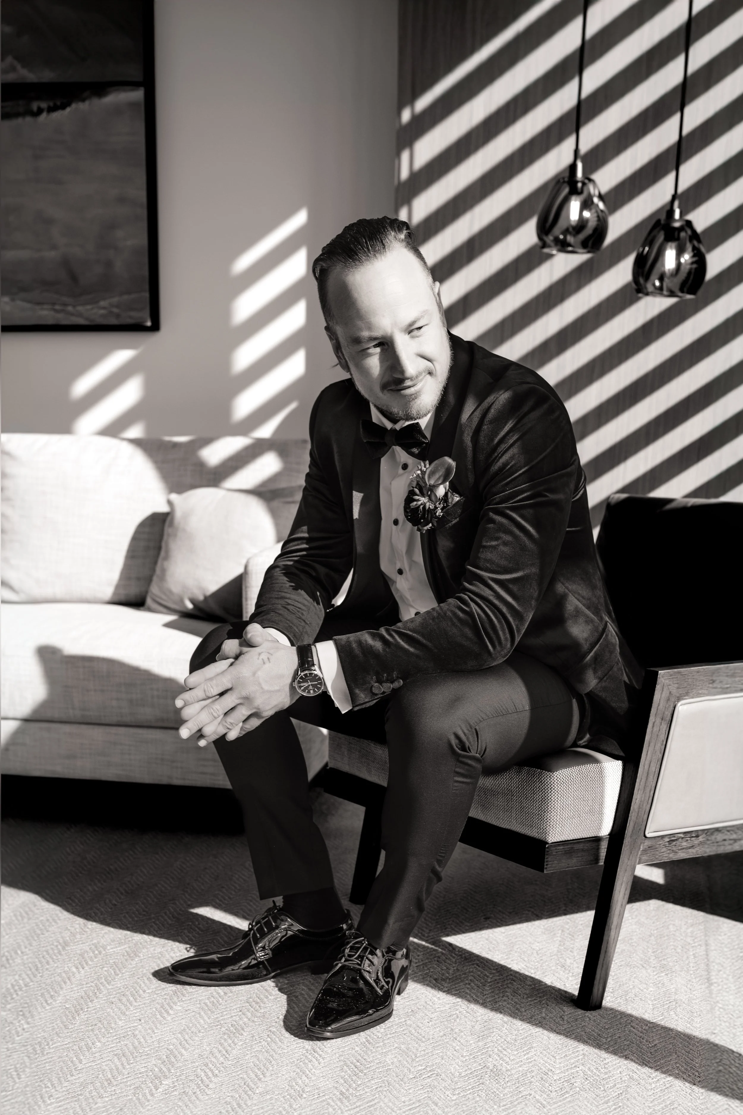 A man in a tuxedo and bowtie sitting on a modern chair, with sunlight streaming through window blinds creating striped shadows on the wall behind him.
