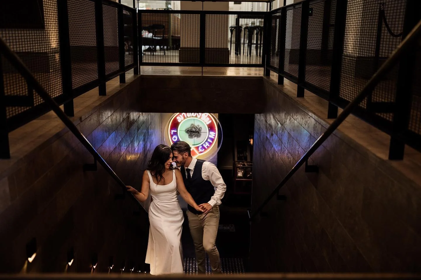 A couple dancing on a staircase in a dimly lit venue with a neon sign in the background.