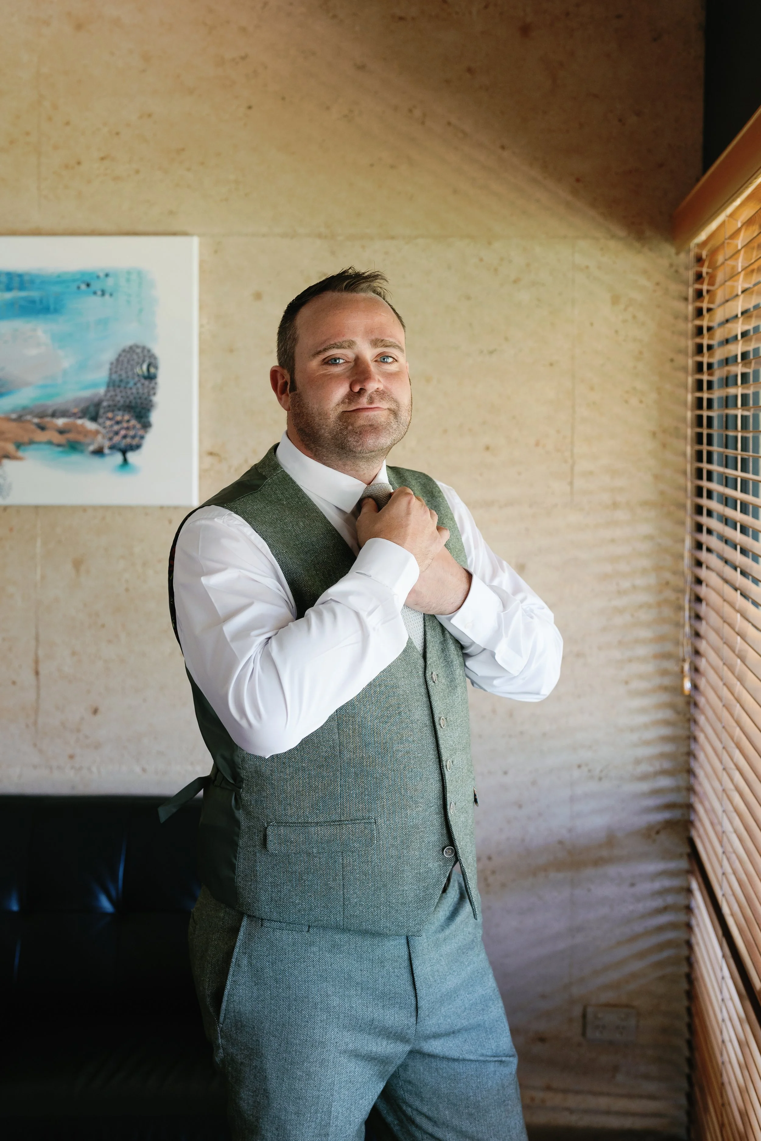A man adjusts his tie while standing in a room with beige walls and wooden blinds, wearing a white shirt and gray vest and pants.