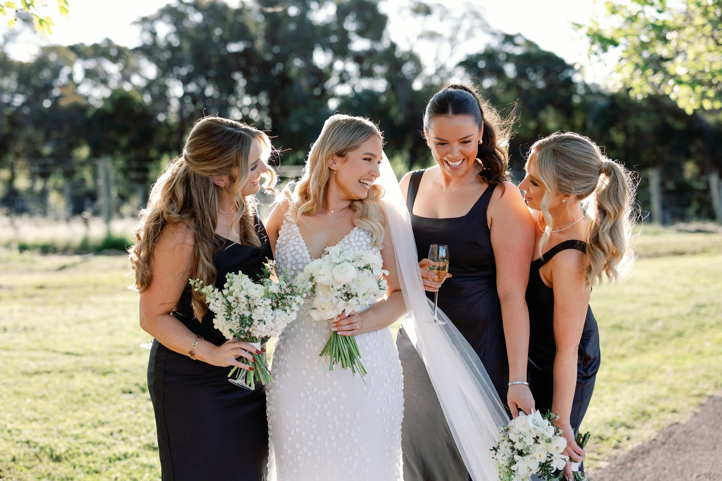 A group of five women, including a bride in a white wedding dress, standing outdoors on a sunny day. The bride is holding a bouquet of white flowers, and the other women are dressed in black dresses, holding bouquets and a glass of champagne, smiling