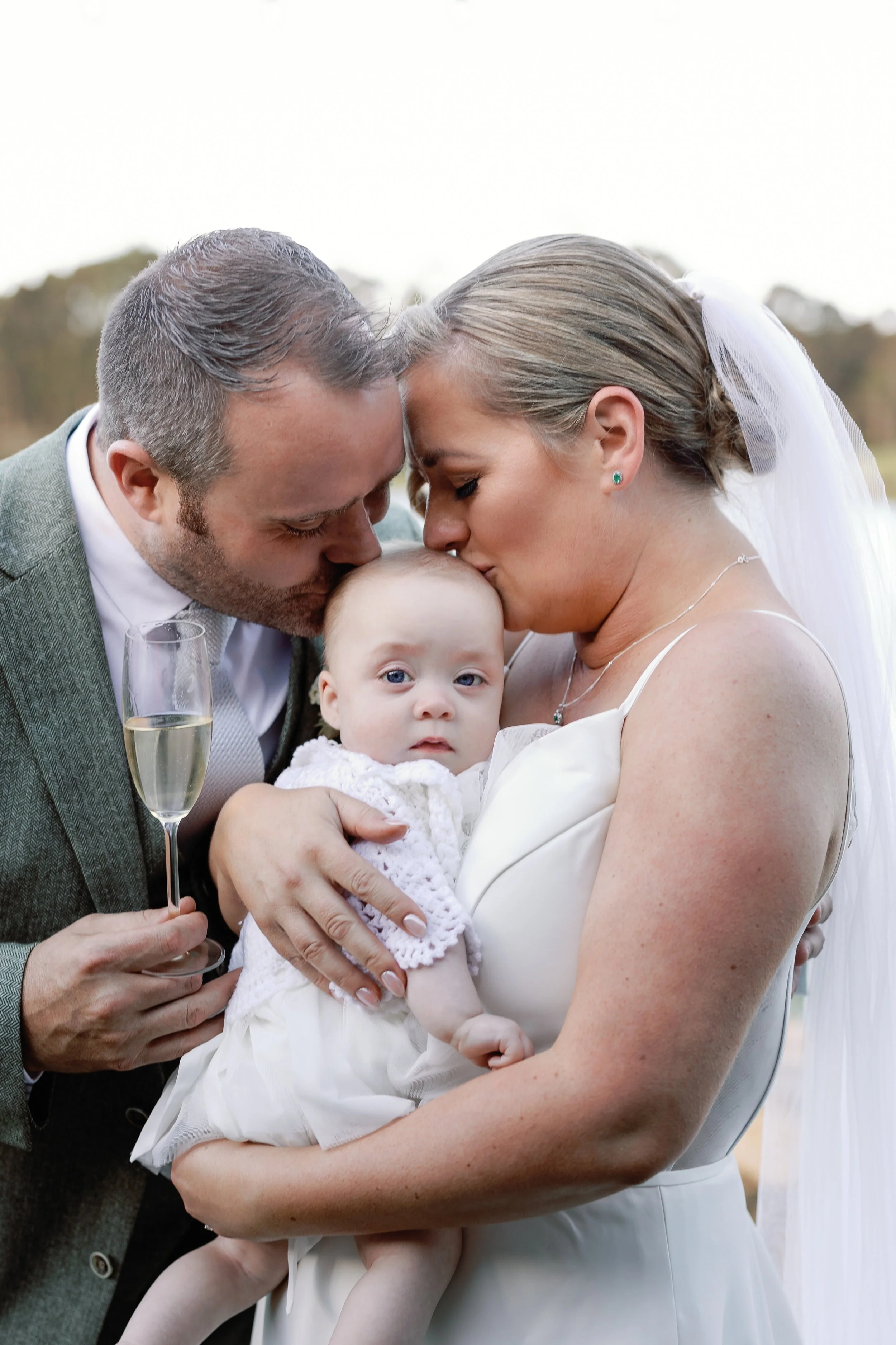 A bride and groom sharing a kiss on their wedding day while holding a baby girl, with the groom holding a glass of champagne.