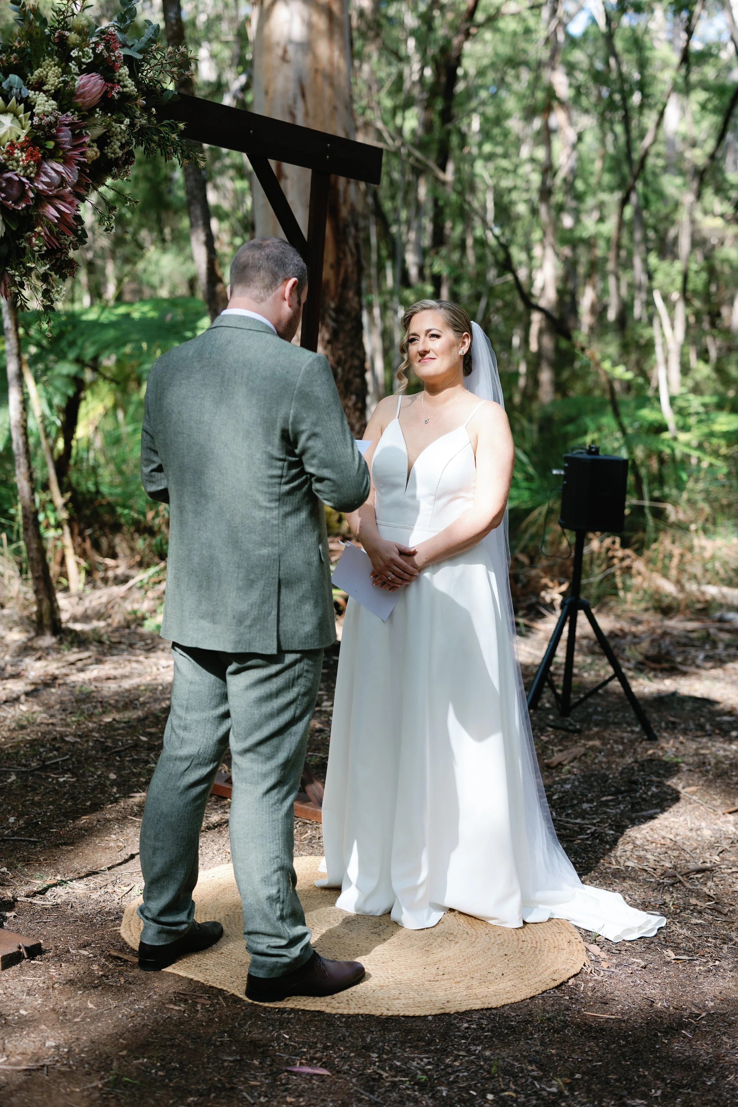 A bride and groom exchanging vows during an outdoor wedding ceremony in a wooded area, with a floral arch overhead and a speaker on a stand nearby.