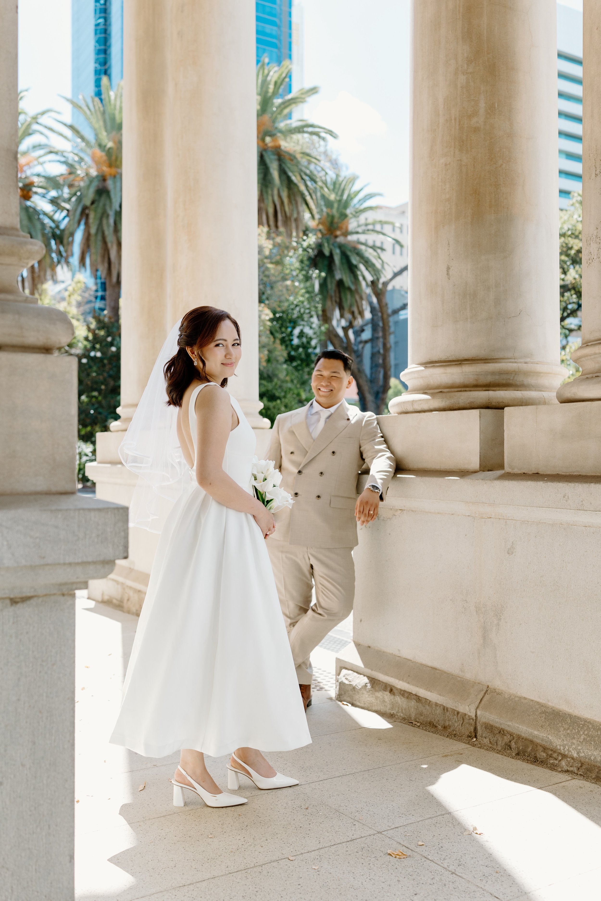 A bride and groom in wedding attire standing outdoors near large stone columns, with palm trees and skyscrapers in the background, under bright sunlight at The State Buildings Perth