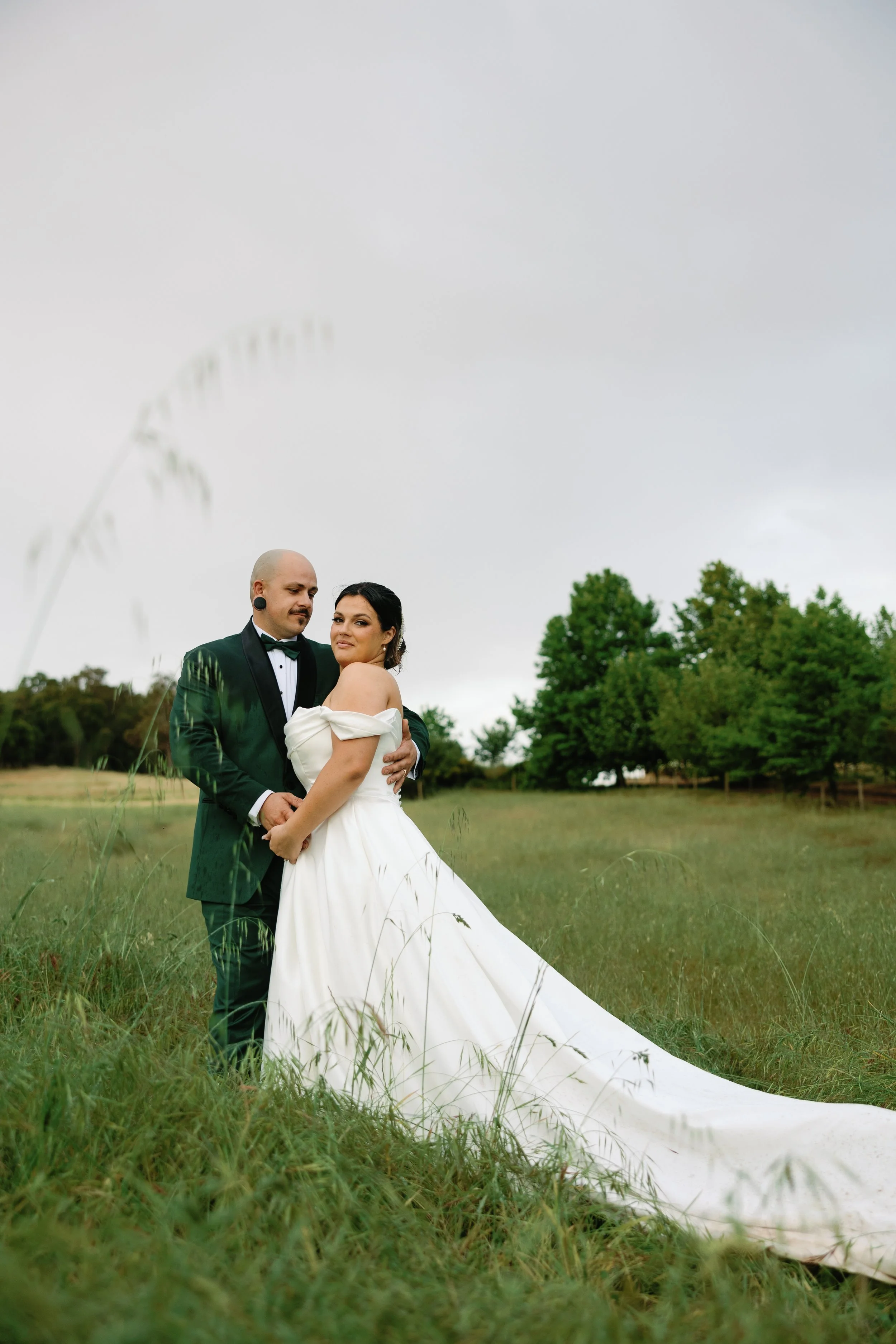Couple in wedding attire standing in a grassy field with trees in the background, on a cloudy day.