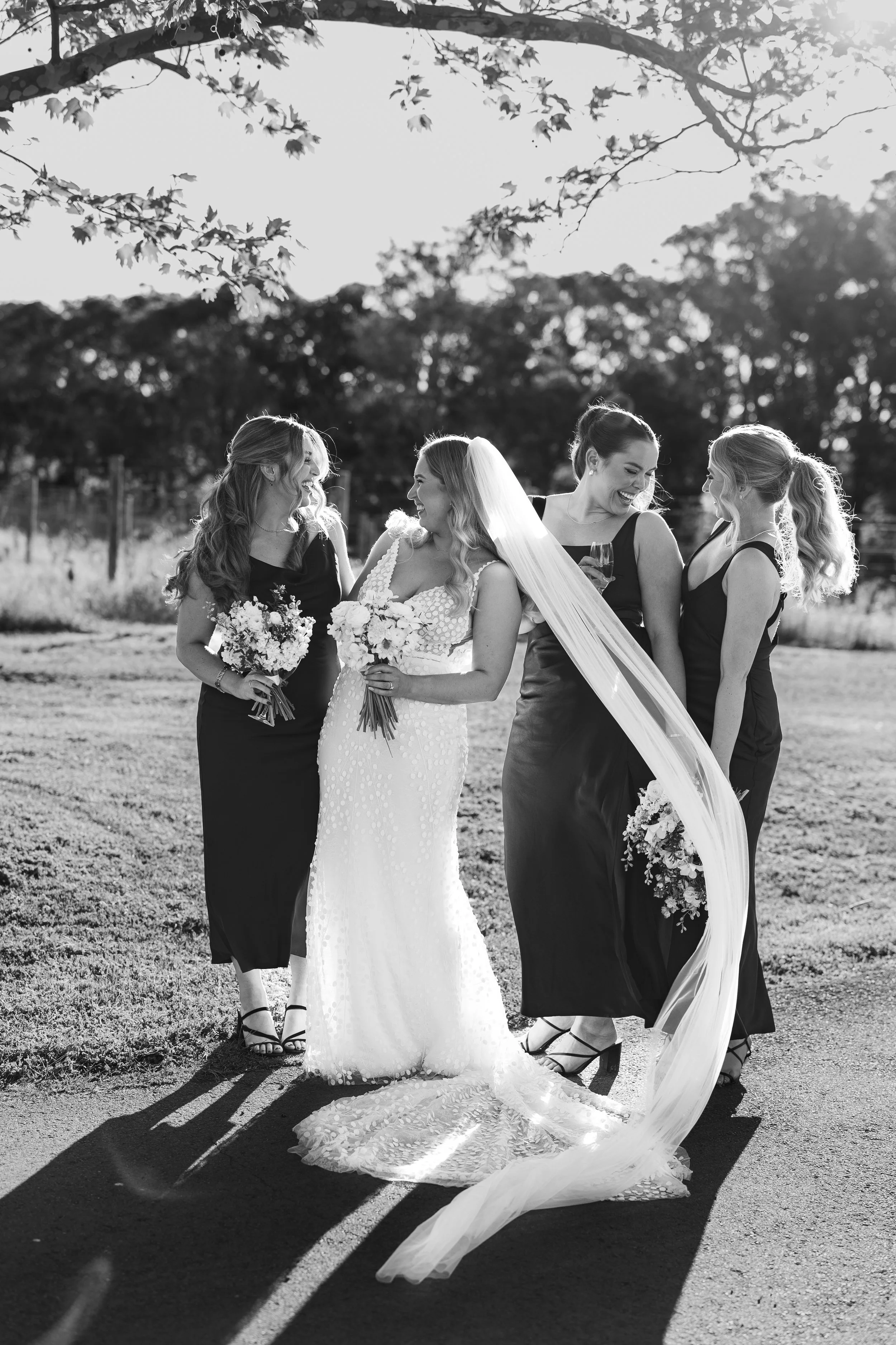 Black and white photo of a bride in a wedding dress with three bridesmaids, all holding bouquets, outdoors with trees and sunlight in the background, smiling and laughing.