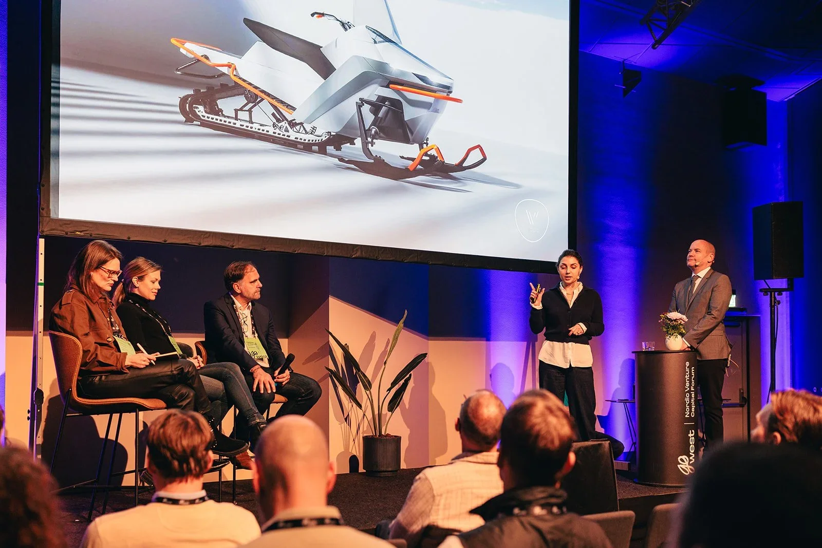 People giving a presentation about a snowmobile on stage at a conference, with an audience watching.