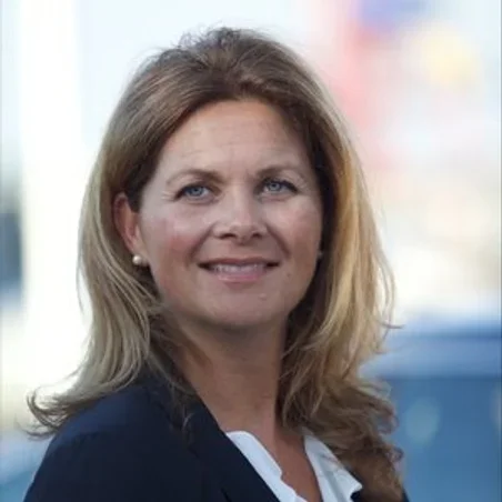 Portrait of a woman with blond hair, wearing a dark blazer and white shirt, smiling outdoors.