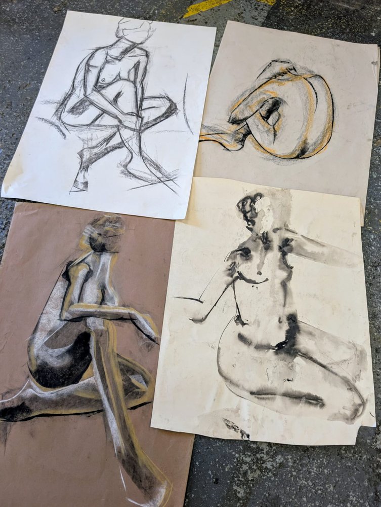 Four life drawing sketches of female nudes on paper, displayed on a workshop floor.