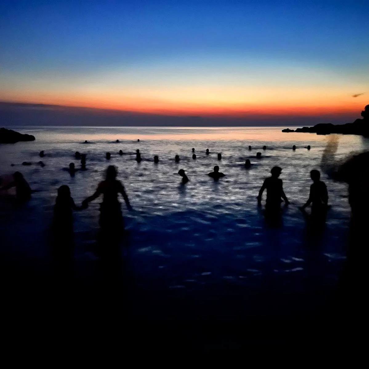 a night time image of sillouette of people in the water with the sun disappearing