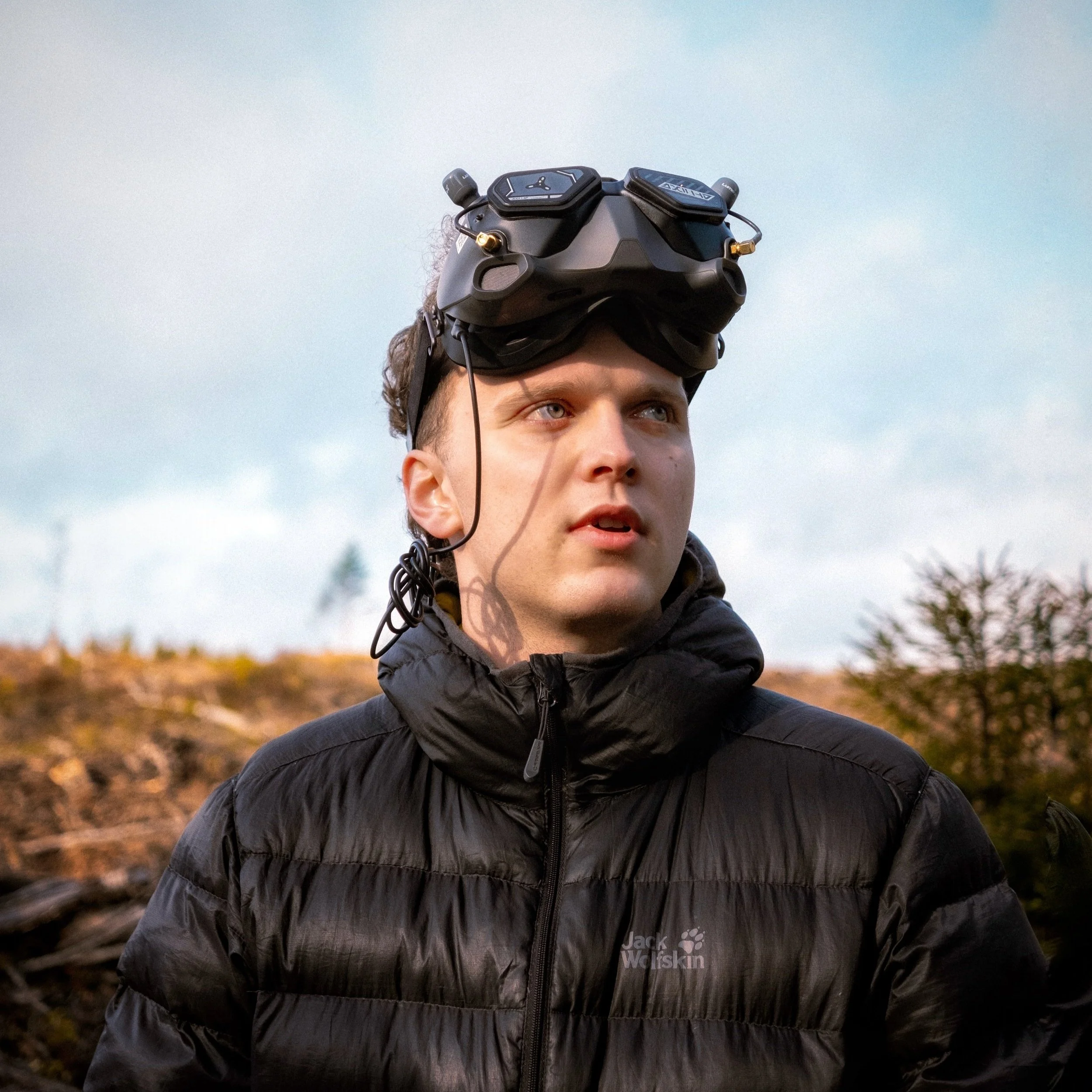 Charlie Ord wearing a black puffy jacket and goggles with a drone controller on his head outdoors on a cloudy day.