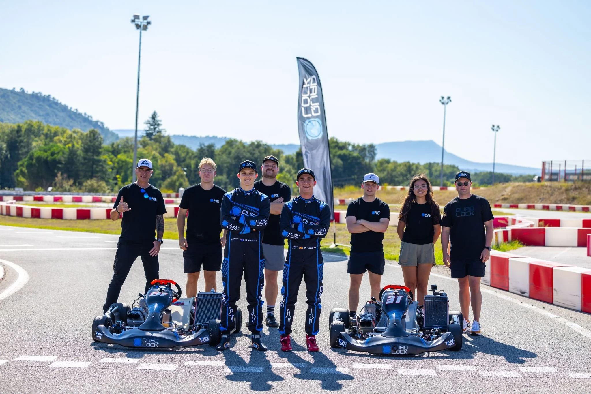 Group of eight people, including race car drivers and team members, standing on a race track with go-karts and a Quad Lock flag, mountains and trees in the background.