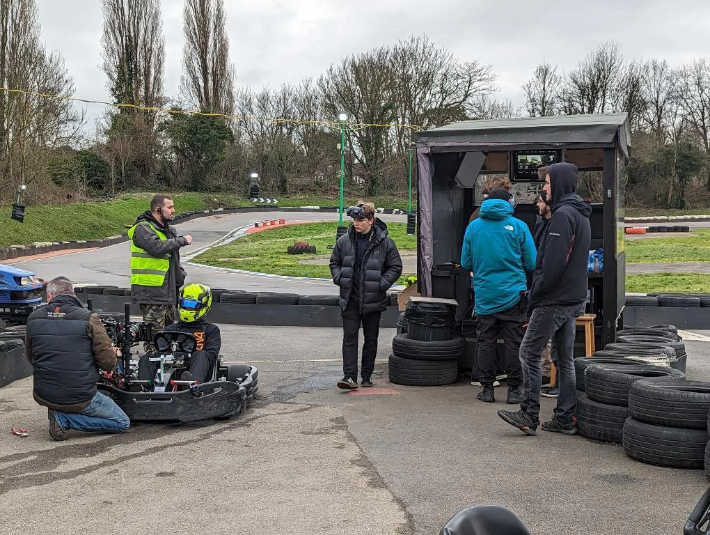 A group of people at a go-kart track, with some crew and a cameraman filming a driver in a go-kart, helmets, and safety gear, on an overcast day.