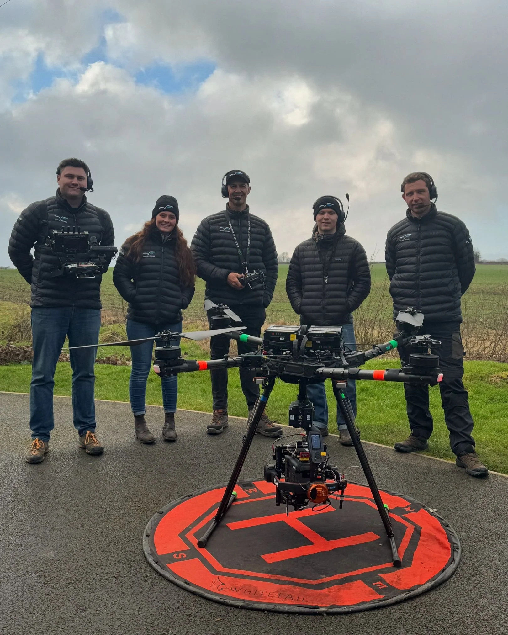 Five people wearing black jackets, standing outdoors on a cloudy day with a large drone and camera equipment on the ground in front of them.