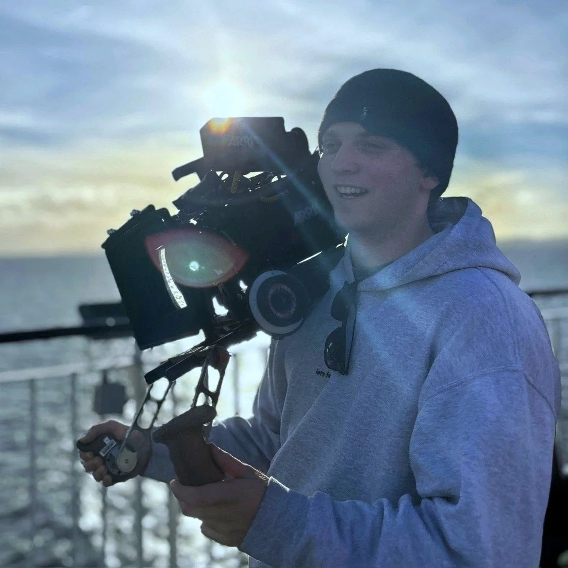 Young man outdoors holding a professional camera with the sun setting or rising behind him, wearing a gray hoodie and black beanie.