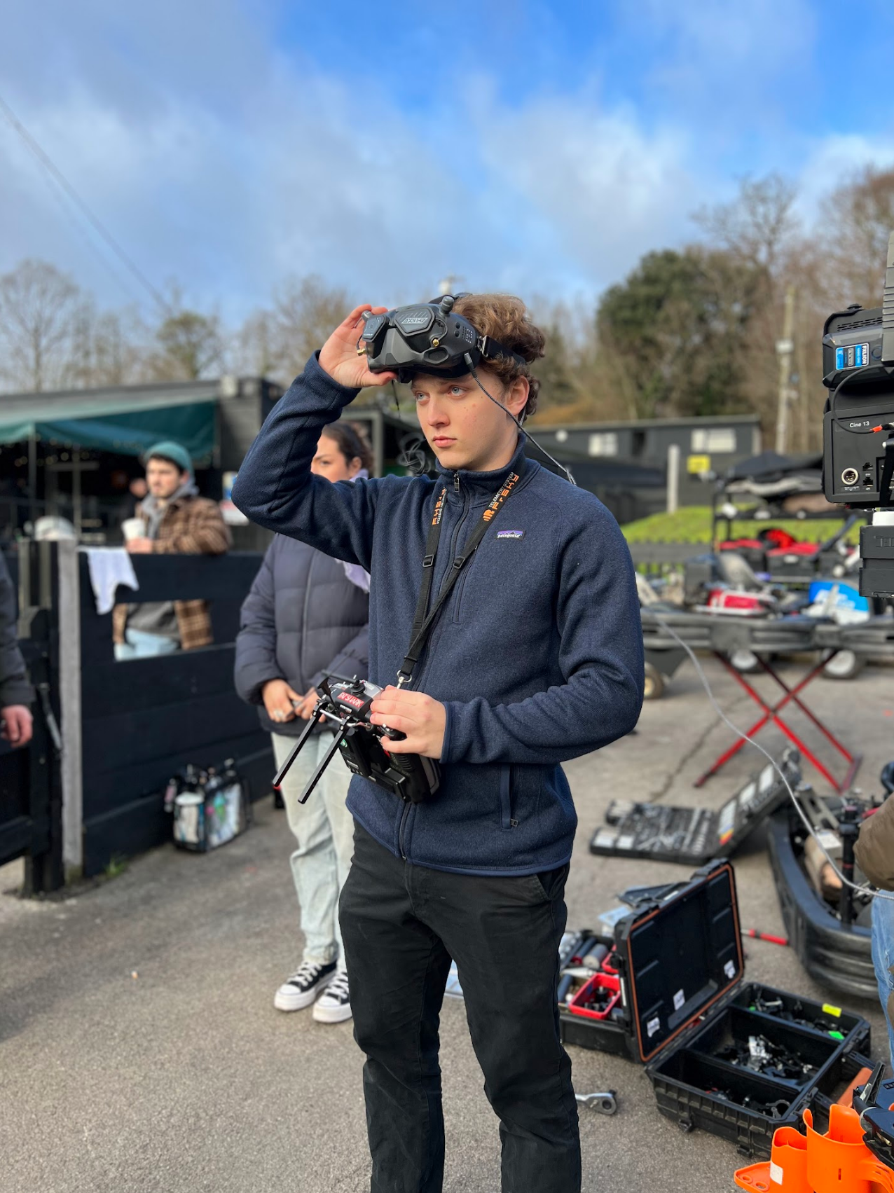Charlie Ord wearing a dark blue fleece jacket and black pants operating a drone controller, holding a virtual reality headset, in an outdoor setting with other people and equipment around.