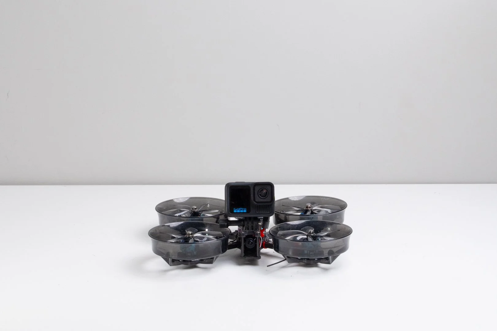 A black quadcopter drone with a GoPro camera mounted on top, resting on a white surface in a room with a light gray wall.