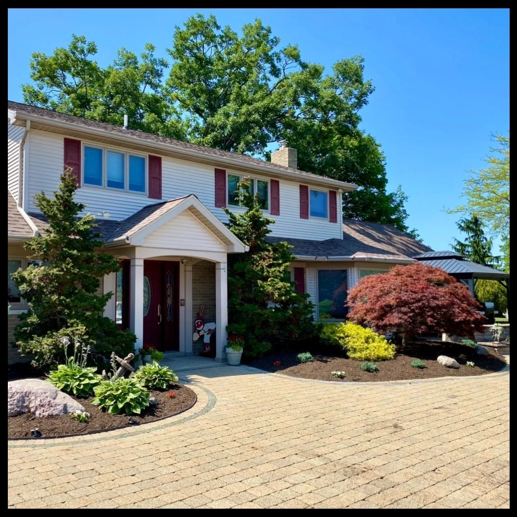 Front view of a two-story house with white siding, red shutters, and a brick chimney, surrounded by landscaped garden with trees, shrubs, and a paved driveway.