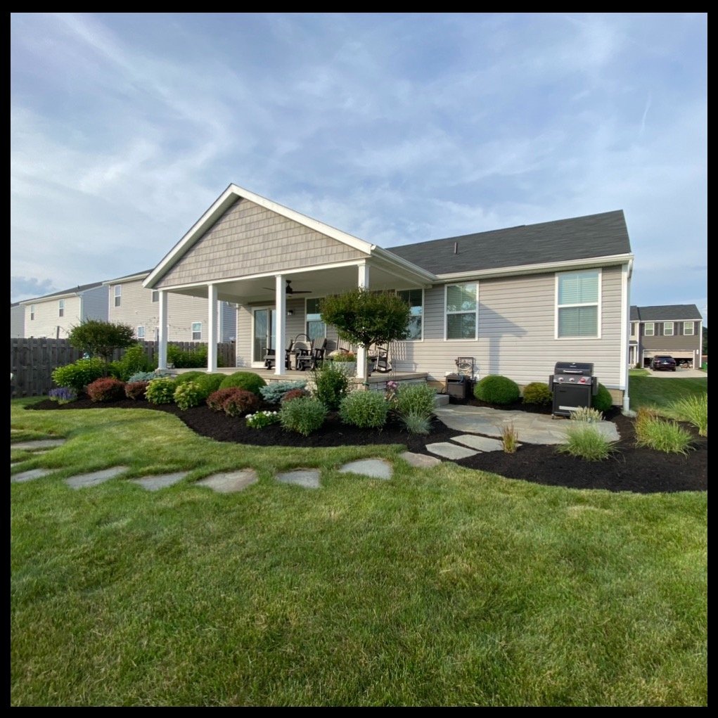 A well-maintained suburban backyard with a house, covered porch, lush green lawn, stepping stones, landscaped flower beds, and outdoor furniture including a grill.