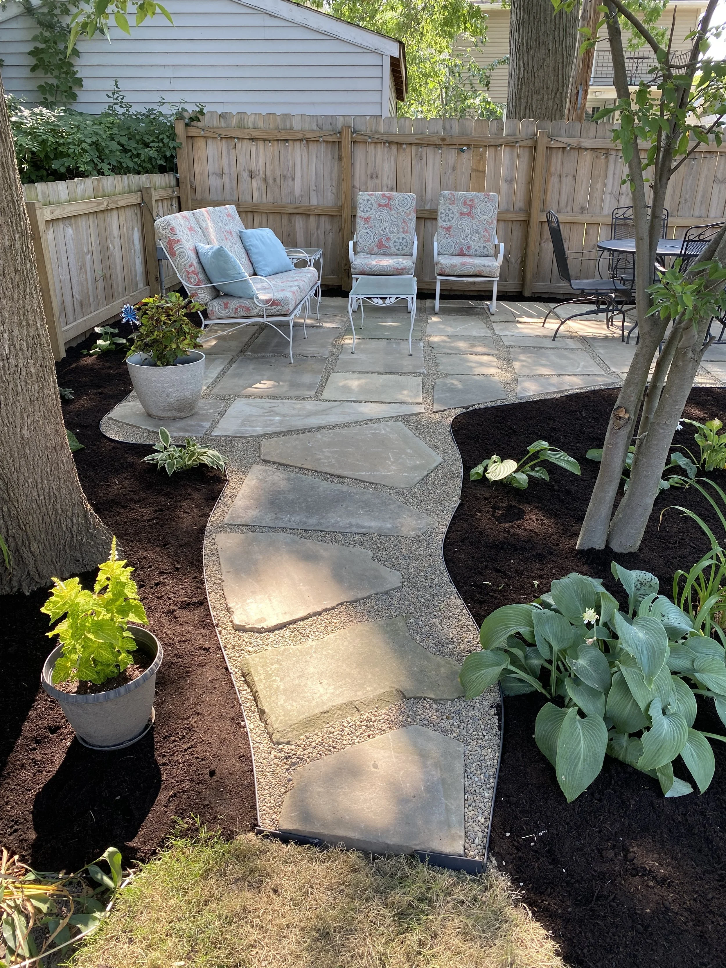 A backyard patio with a curved stone pathway, patio furniture including a cushioned loveseat, two chairs, and a small table, surrounded by flower beds with plants and mulch, enclosed by a wooden fence and trees.