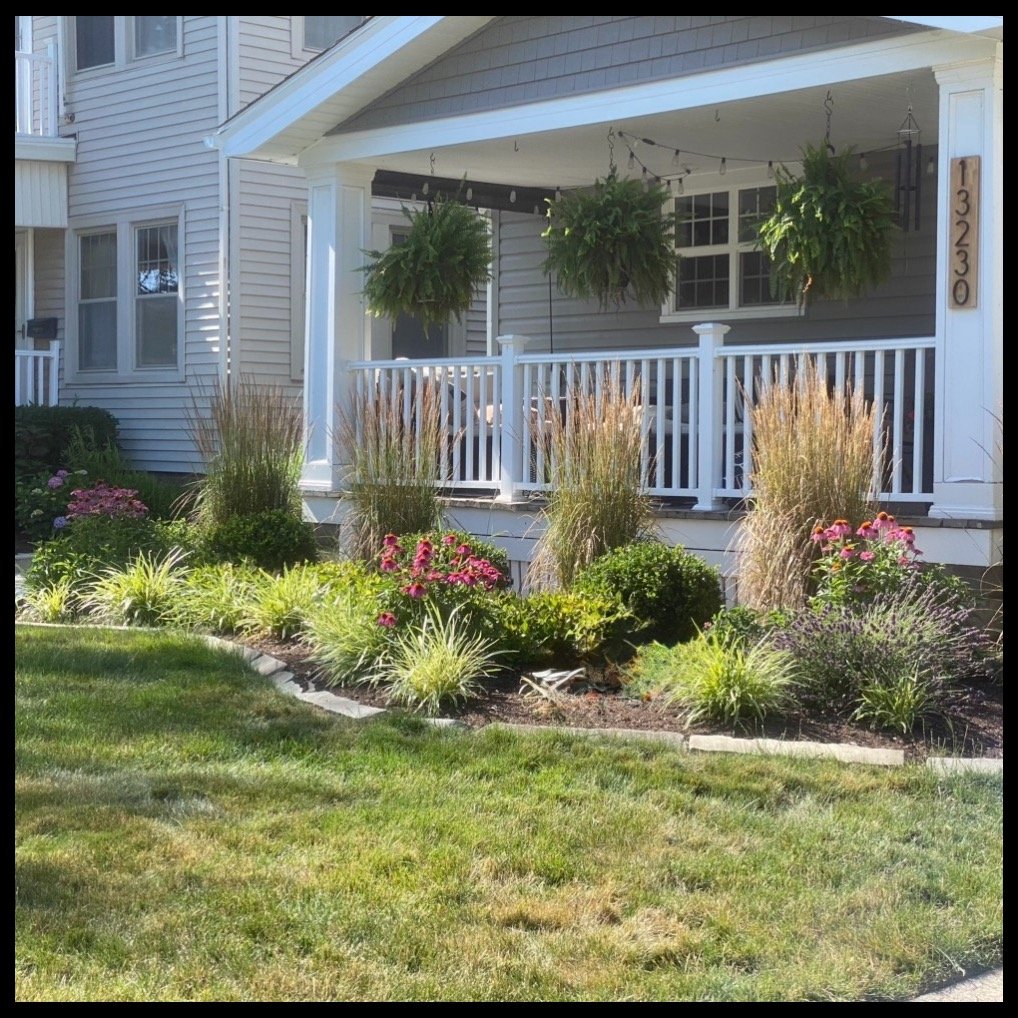 Front porch of a house with hanging potted plants, white railing, and a garden bed with various plants and flowers in front.