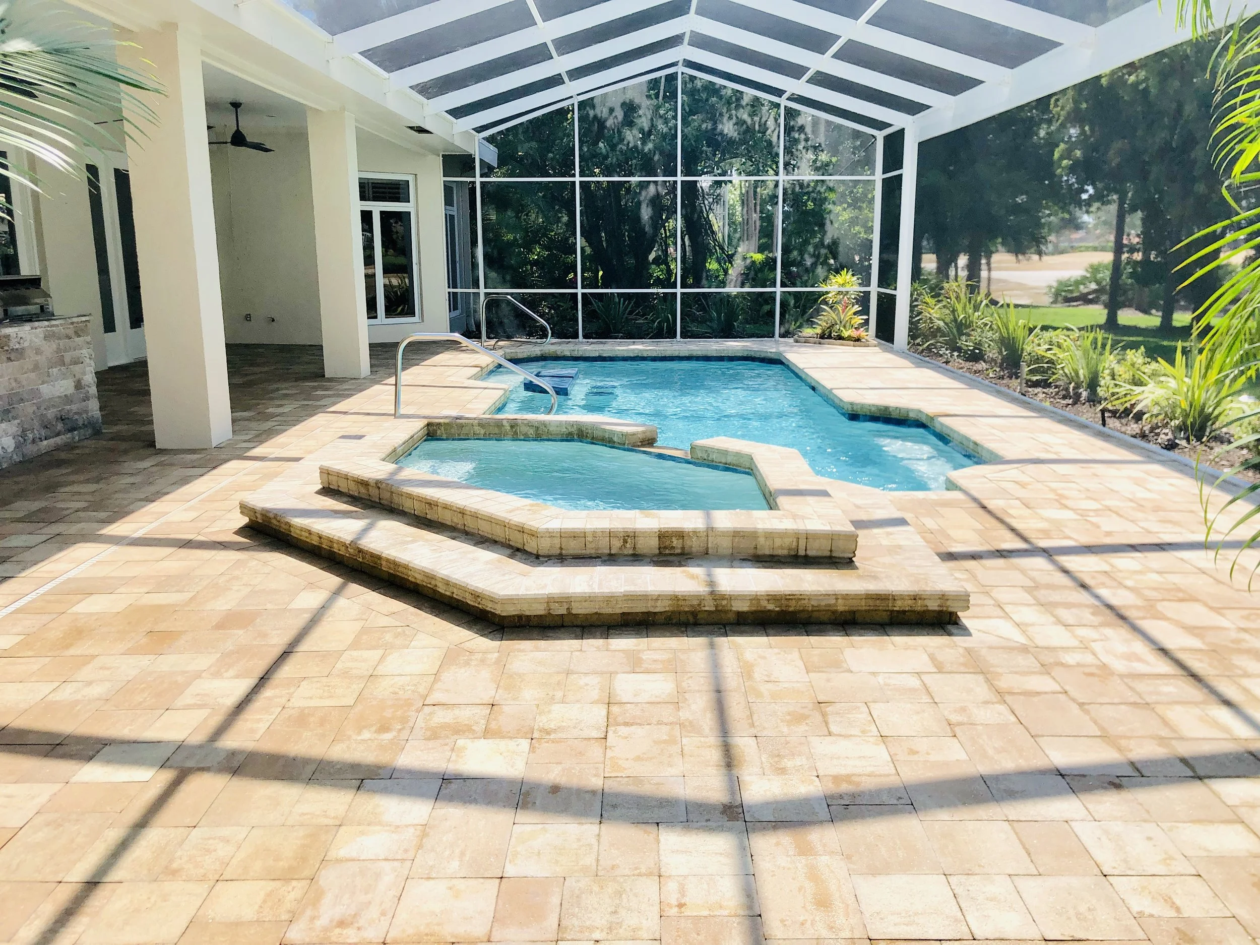 A backyard patio with a small pool and hot tub, surrounded by a screened enclosure, with outdoor plants and trees visible beyond.