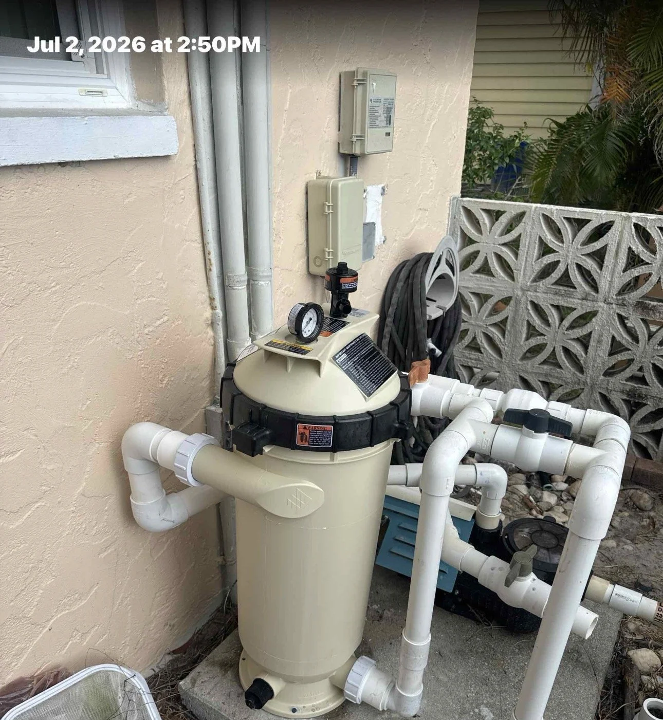 Pool filter and pump system outside near a beige wall with pipes and electrical boxes, surrounded by a decorative concrete block wall and some greenery.