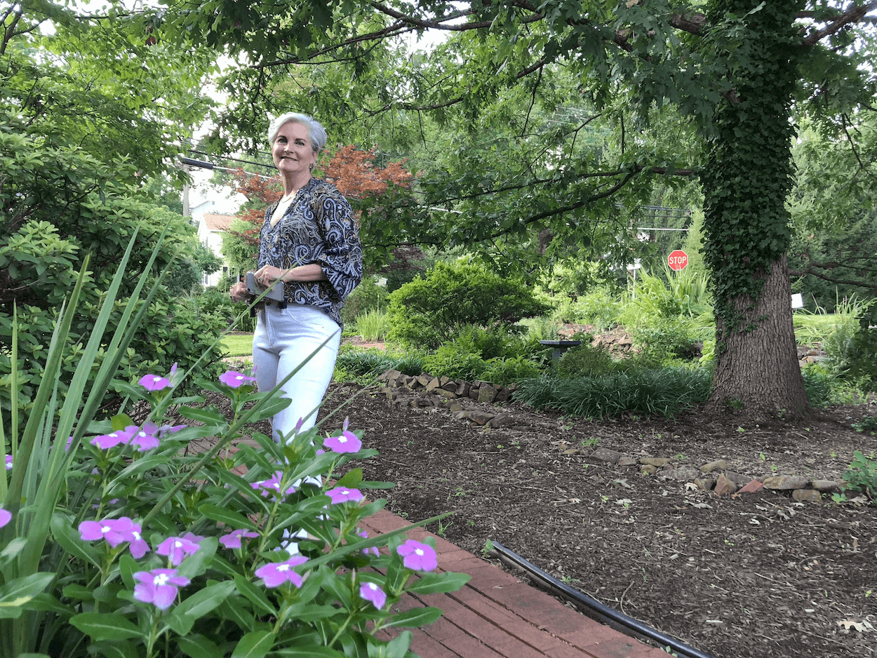 Christina stands in a garden surrounded by plants and flowers. She smiles at the camera