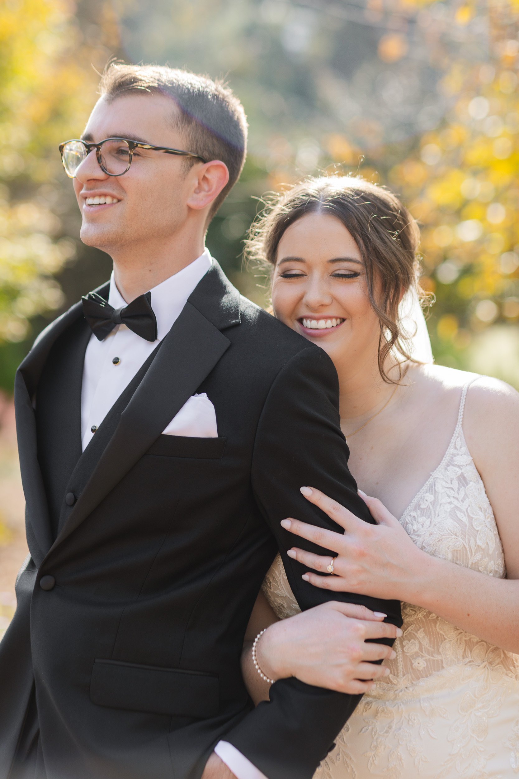 A smiling bride hugging a groom from behind outdoors during daytime, with the groom in a black tuxedo and glasses, and the bride in a white lace wedding dress during intimate wedding in Travelers Rest, SC
