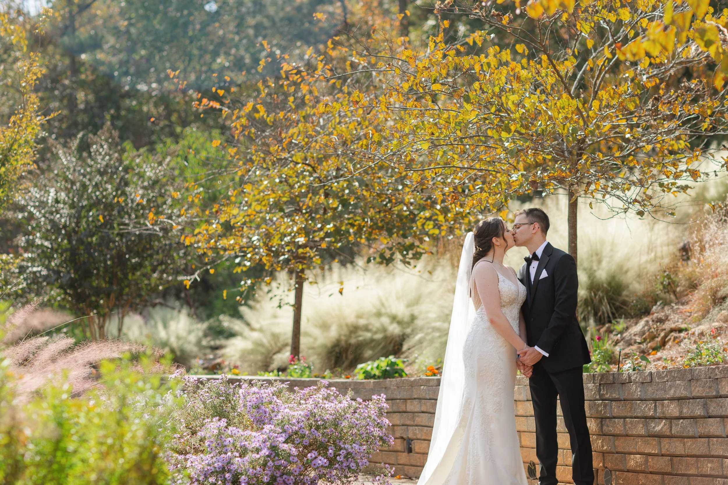 A bride and groom share a kiss outdoors during their wedding in a garden with trees and colorful flowers during intimate wedding in Greenville, SC
