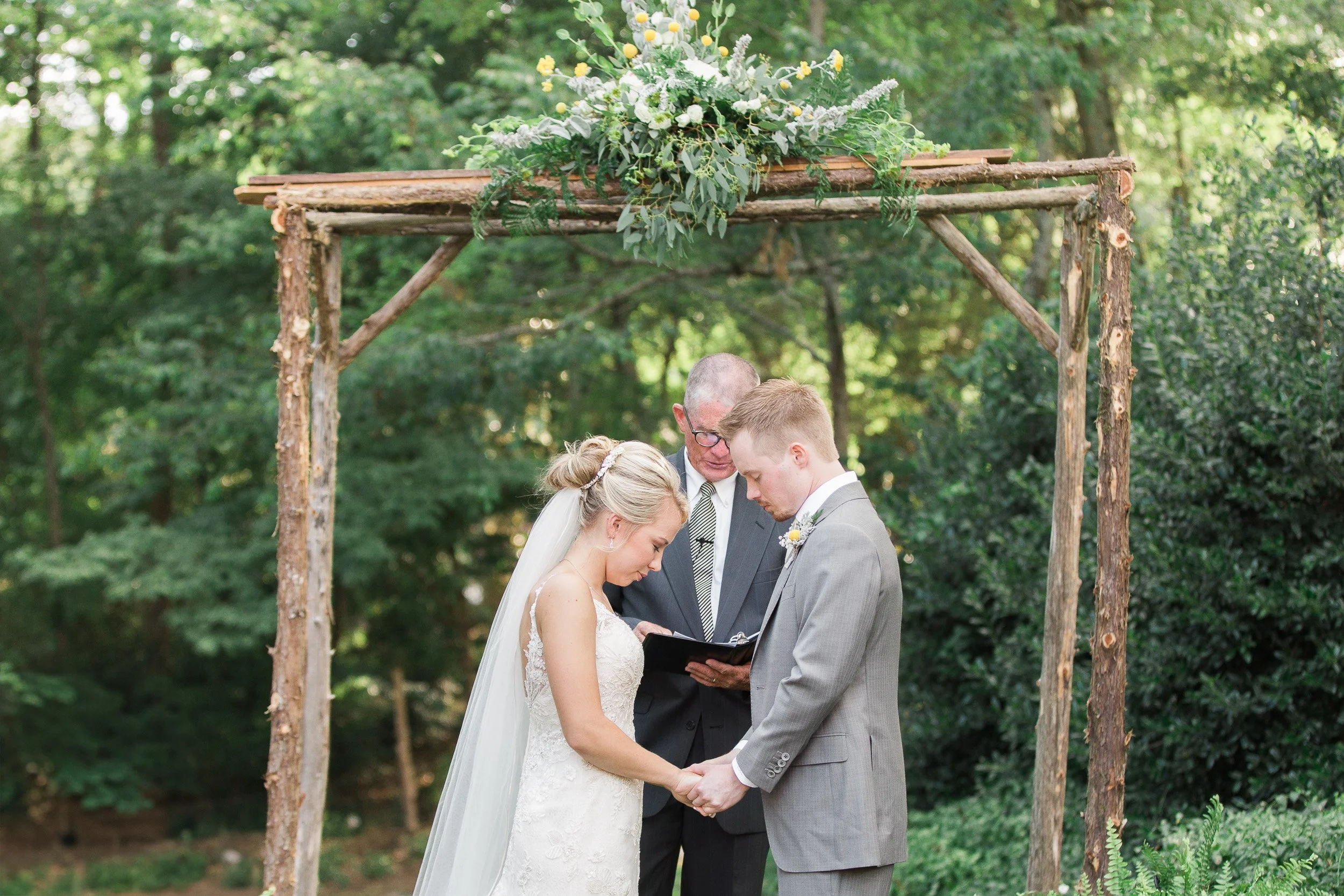 A bride and groom standing under a rustic wooden arch with a floral arrangement, holding hands and facing each other during a wedding ceremony outdoors surrounded by green trees during intimate wedding at Lake Keowee in South Carolina