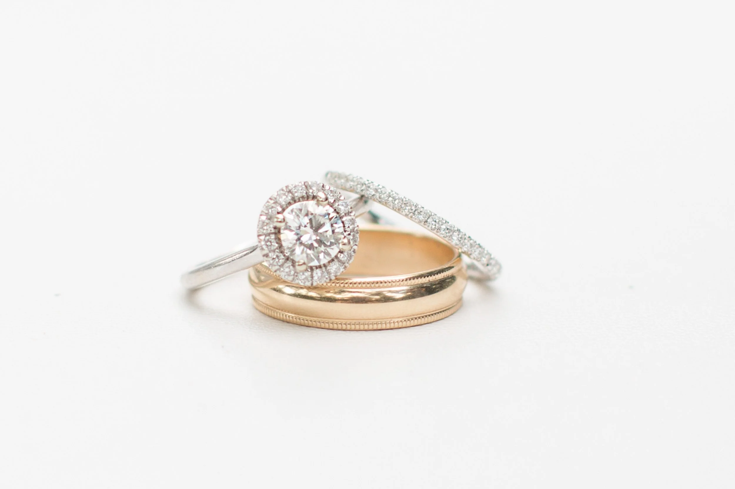 Close-up of three rings, a diamond engagement ring, a wedding band, and an anniversary band, resting on a marble surface with a white background during intimate wedding in Central, SC