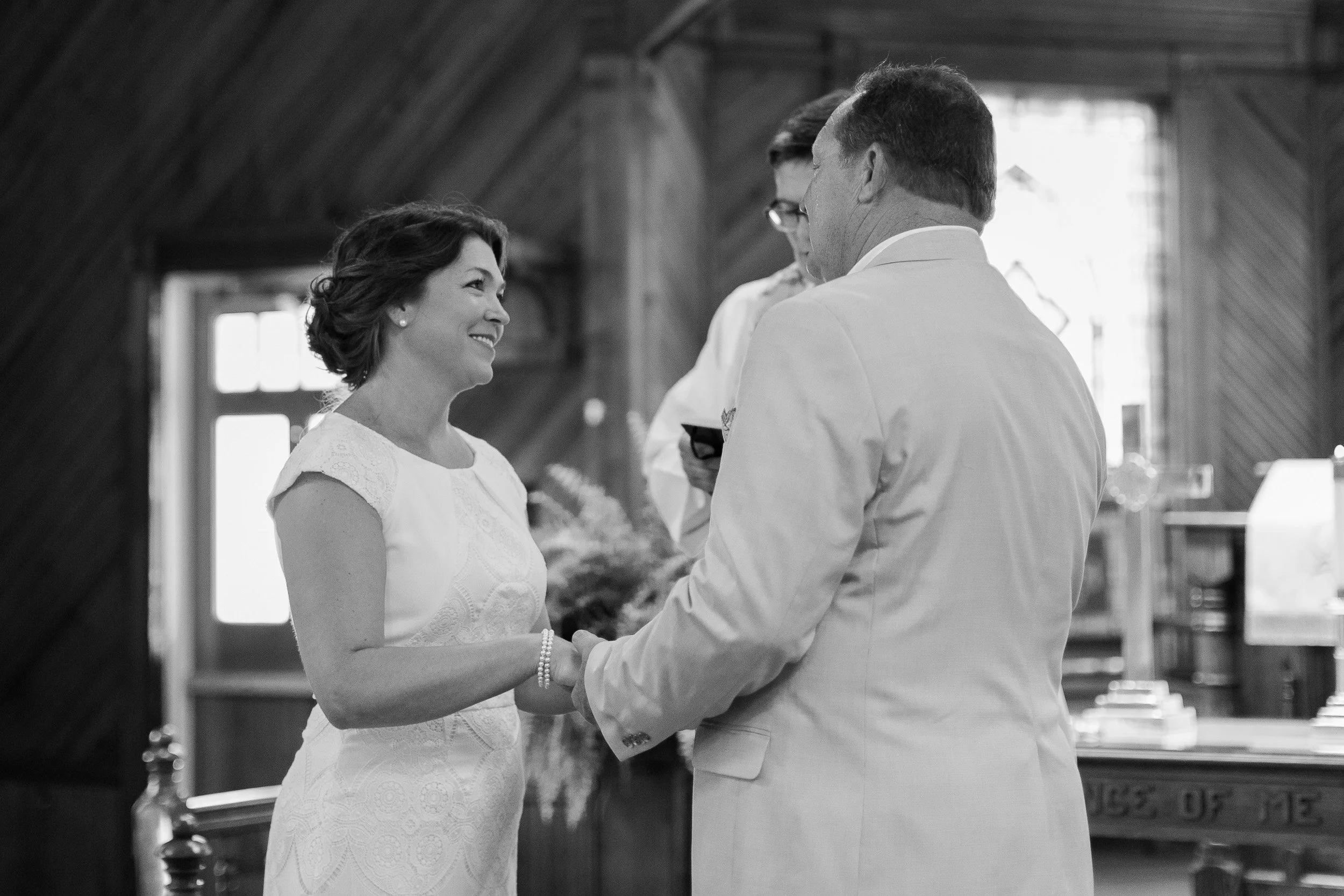 A woman in a white dress and pearl bracelet smiling during a wedding ceremony, holding hands with a man in a white suit inside a wooden church, with officiant in the background in Clemson, SC