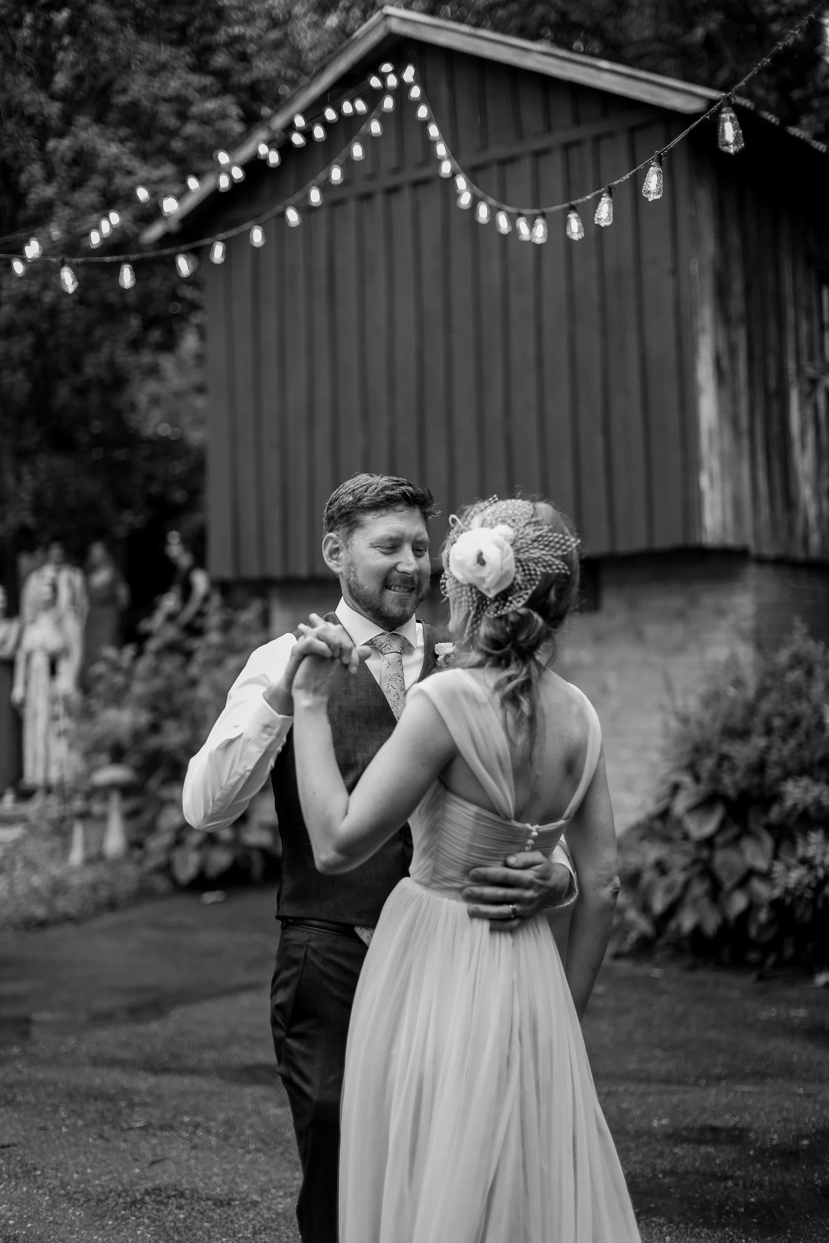 A black and white photo of a man and woman dancing outdoors with string lights overhead and a barn in the background.