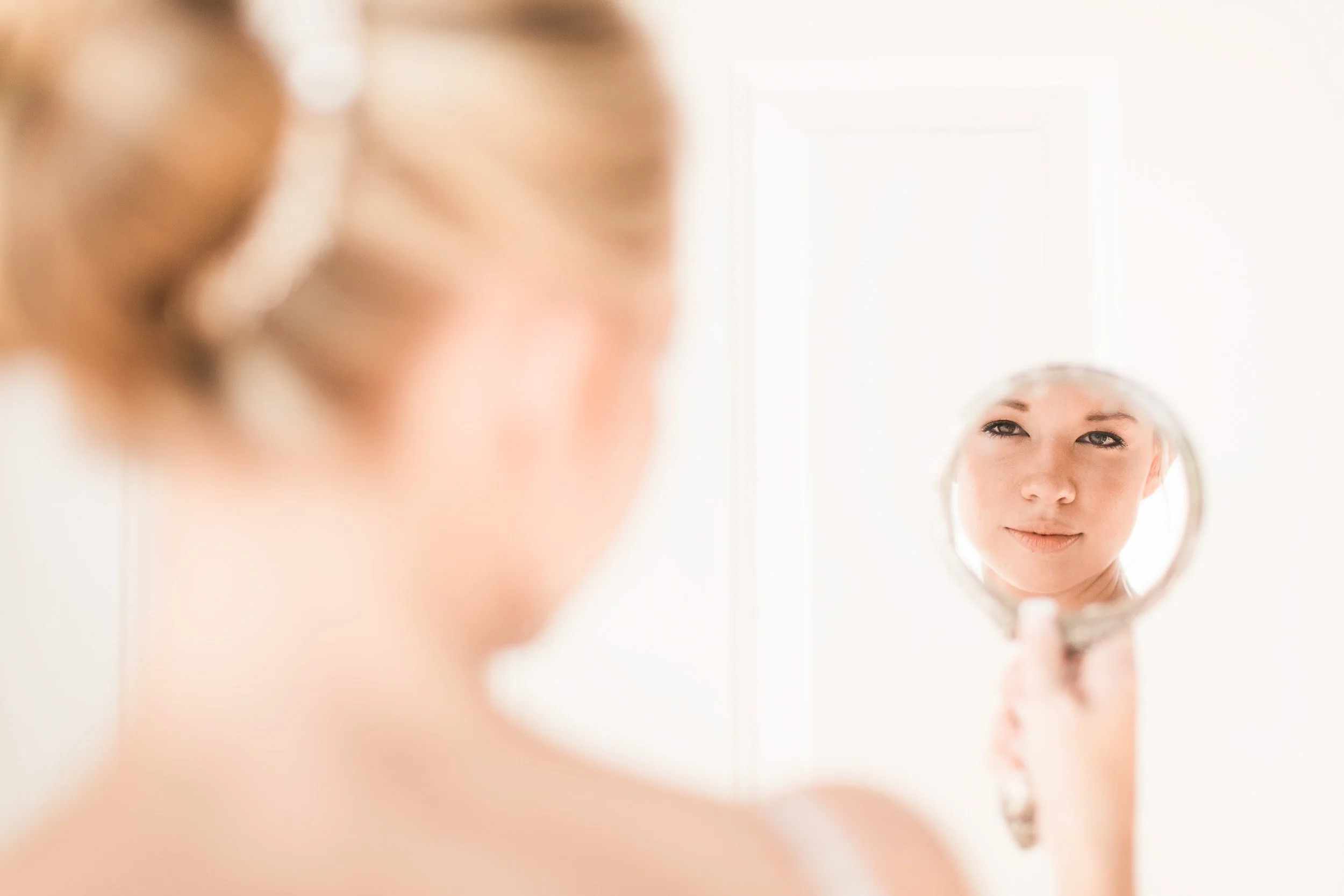 A bride holding a small mirror shows their face reflected in it. The brides face is visible in the mirror, while the rest of the person is blurred and out of focus during backyard wedding in Clemson, SC